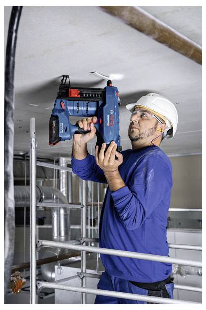 Un ouvrier du bâtiment portant une tenue bleue et un casque de chantier utilise un outil électrique Bosch pour travailler sur le plafond, debout sur un échafaudage dans un environnement intérieur.