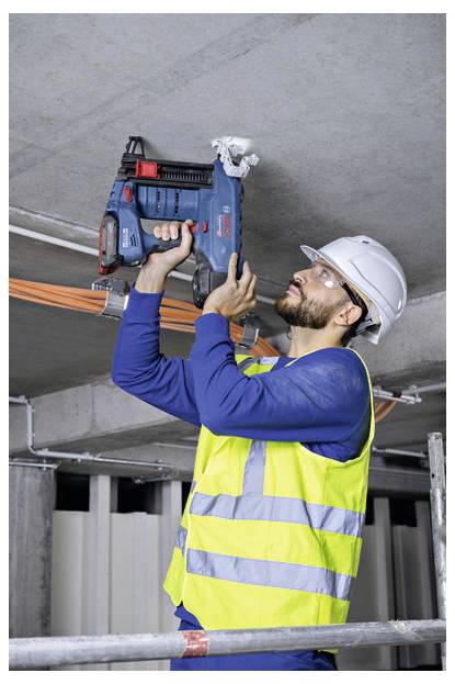 Un ouvrier du bâtiment portant un casque de chantier et un gilet de sécurité utilise un outil électrique pour fixer des câbles au plafond d'un bâtiment en cours de construction.