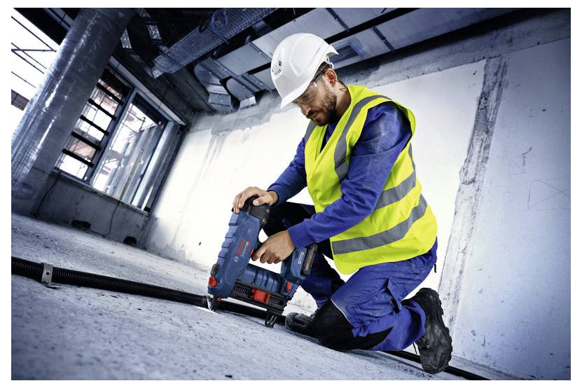 Un ouvrier du bâtiment portant un gilet de sécurité et un casque utilise un outil électrique sur le sol d'un bâtiment en construction.