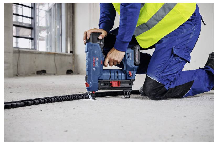 Un ouvrier du bâtiment portant un uniforme bleu et un gilet de sécurité est agenouillé sur un sol en béton, utilisant un outil industriel pour fixer ou attacher des matériaux.