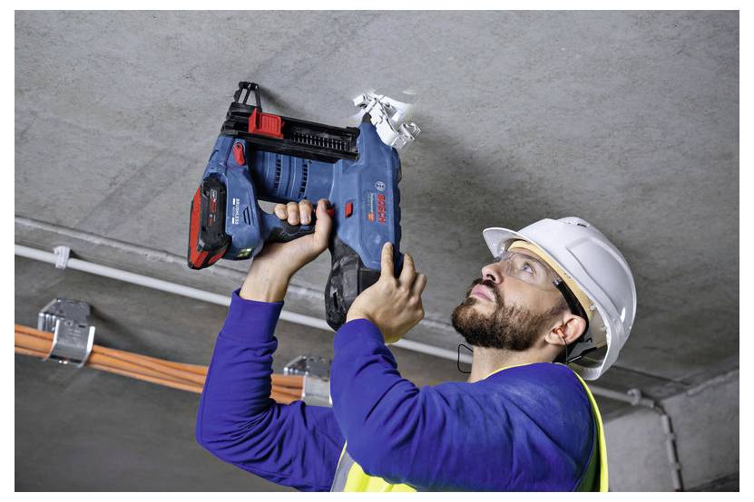 Un ouvrier du bâtiment portant un équipement de sécurité utilise un outil électrique pour percer un plafond en béton, avec des câbles électriques visibles à proximité.