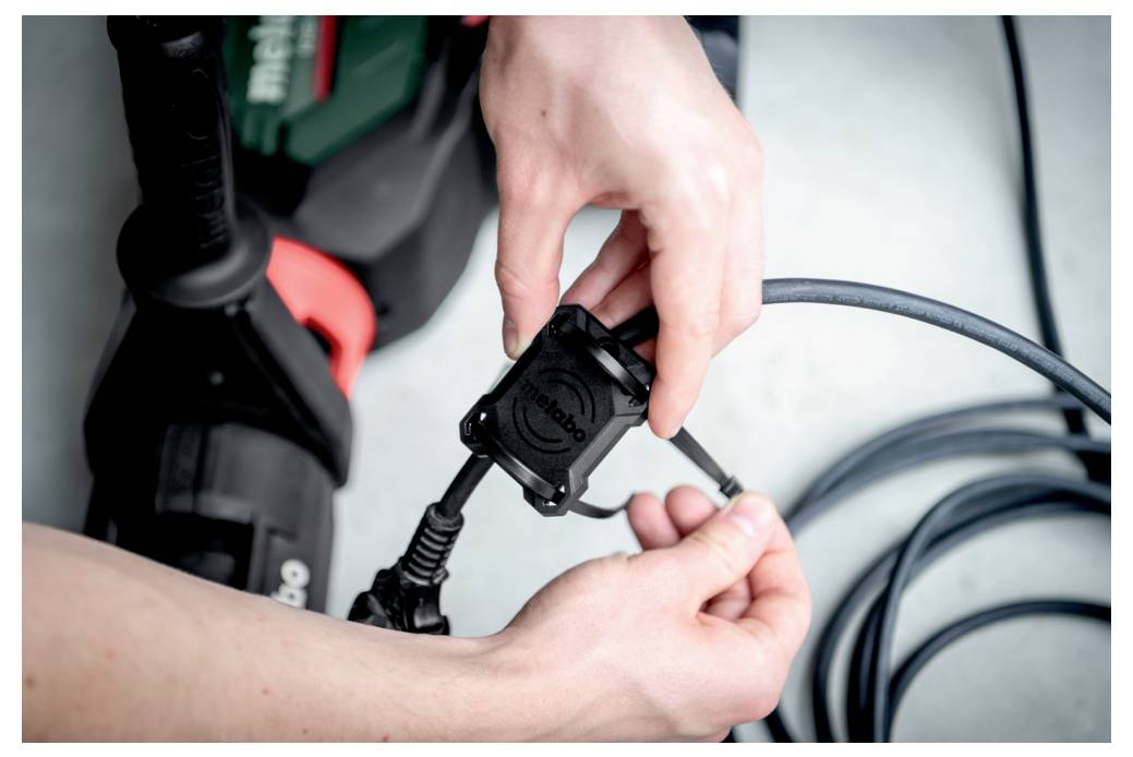 Des mains sont en train de brancher la fiche d'un outil électrique sur une prise de marque Metabo. L'outil et le câble sont positionnés sur une surface claire.
