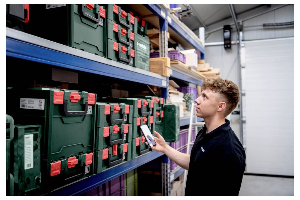 Une personne utilisant un appareil mobile pour scanner des boîtes de rangement vertes avec des loquets rouges sur des étagères dans un environnement d'entrepôt.