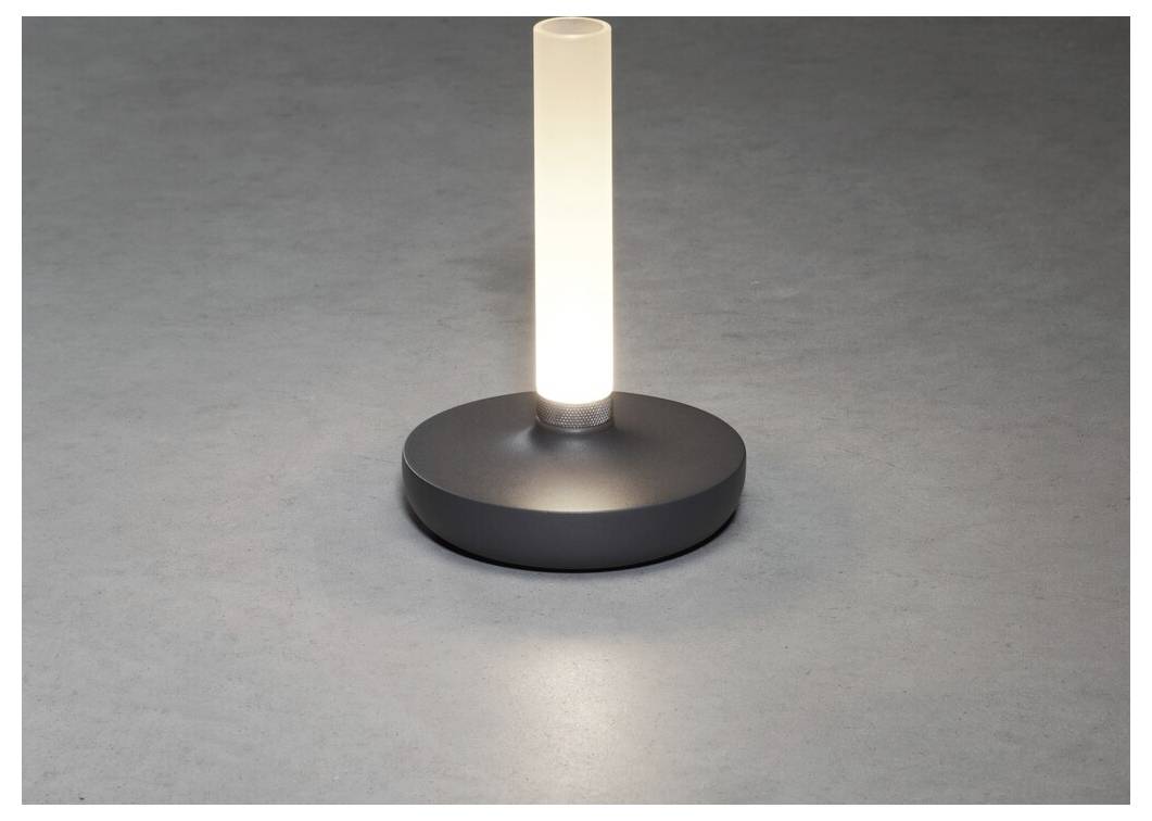 Une petite lampe de table moderne avec un cylindre lumineux et une base ronde émet une lueur douce sur une surface grise.