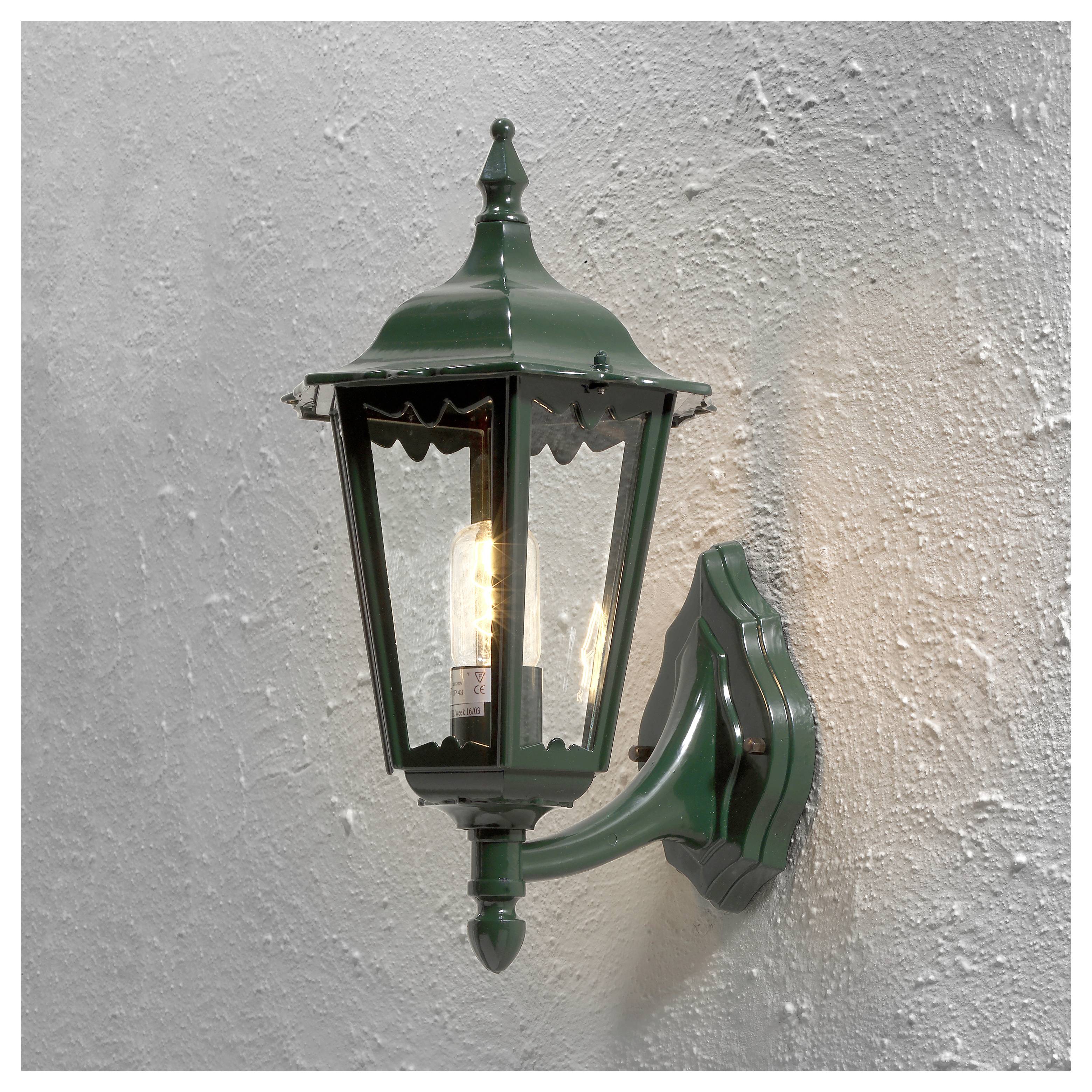 Un lampadaire extérieur de style vintage en vert, monté sur un mur texturé, illuminé par une ampoule brillante à l'intérieur.