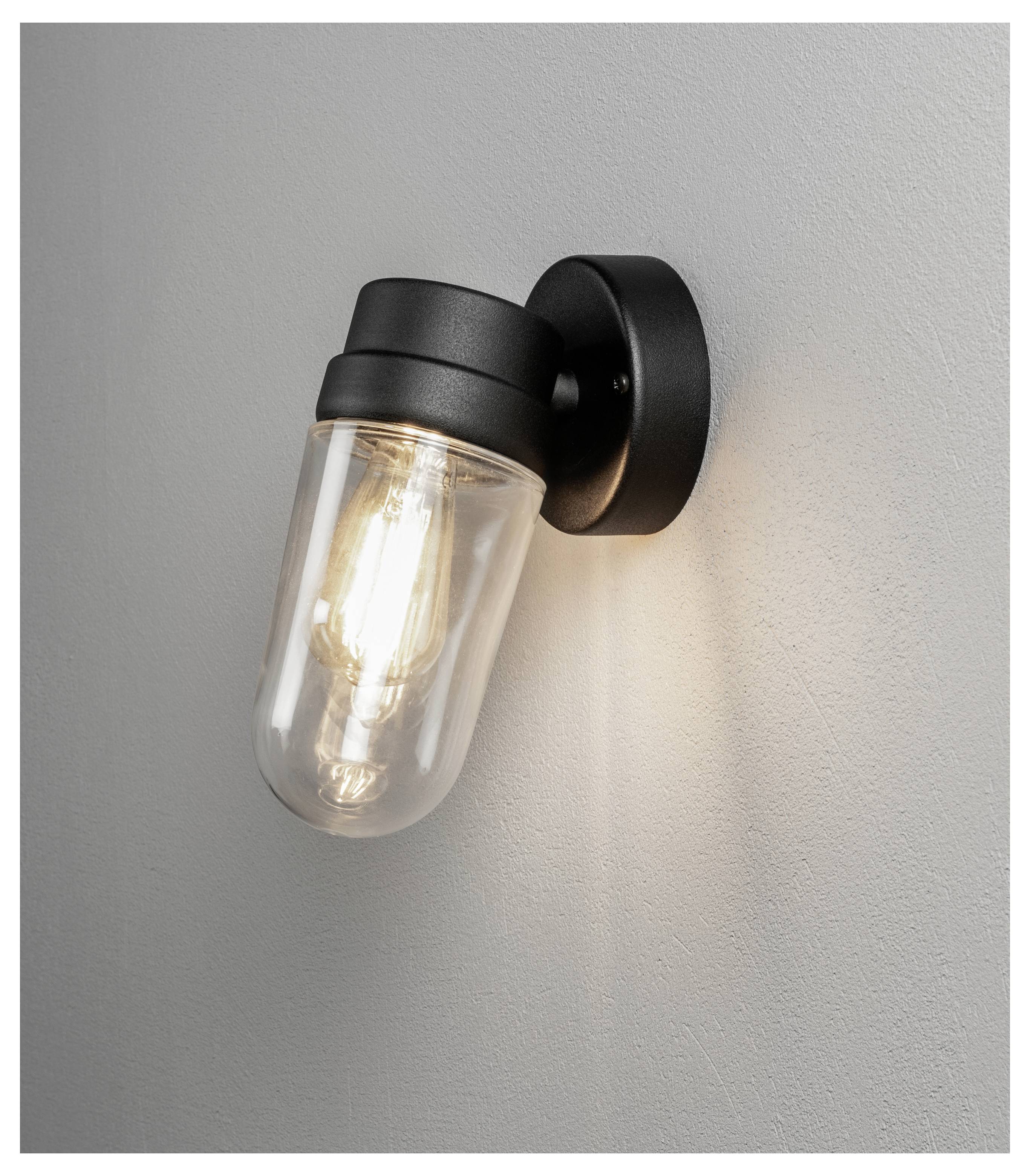 Un applique murale de style industriel avec un abat-jour en verre transparent et une ampoule à filament visible, diffusant une lueur chaleureuse contre un mur gris.
