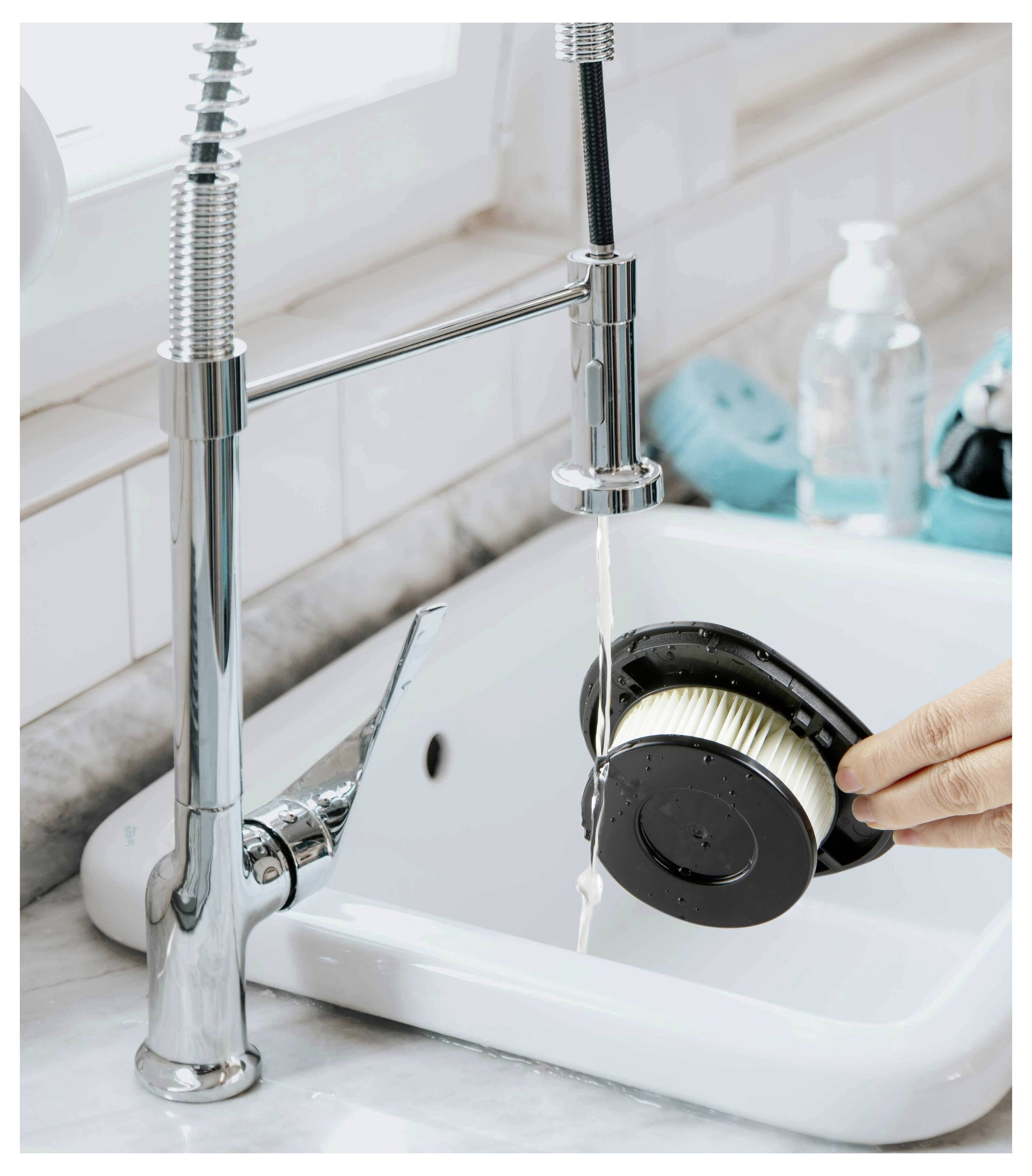 Une personne rince un filtre d'aspirateur sous un robinet de cuisine, avec des produits de nettoyage visibles en arrière-plan.