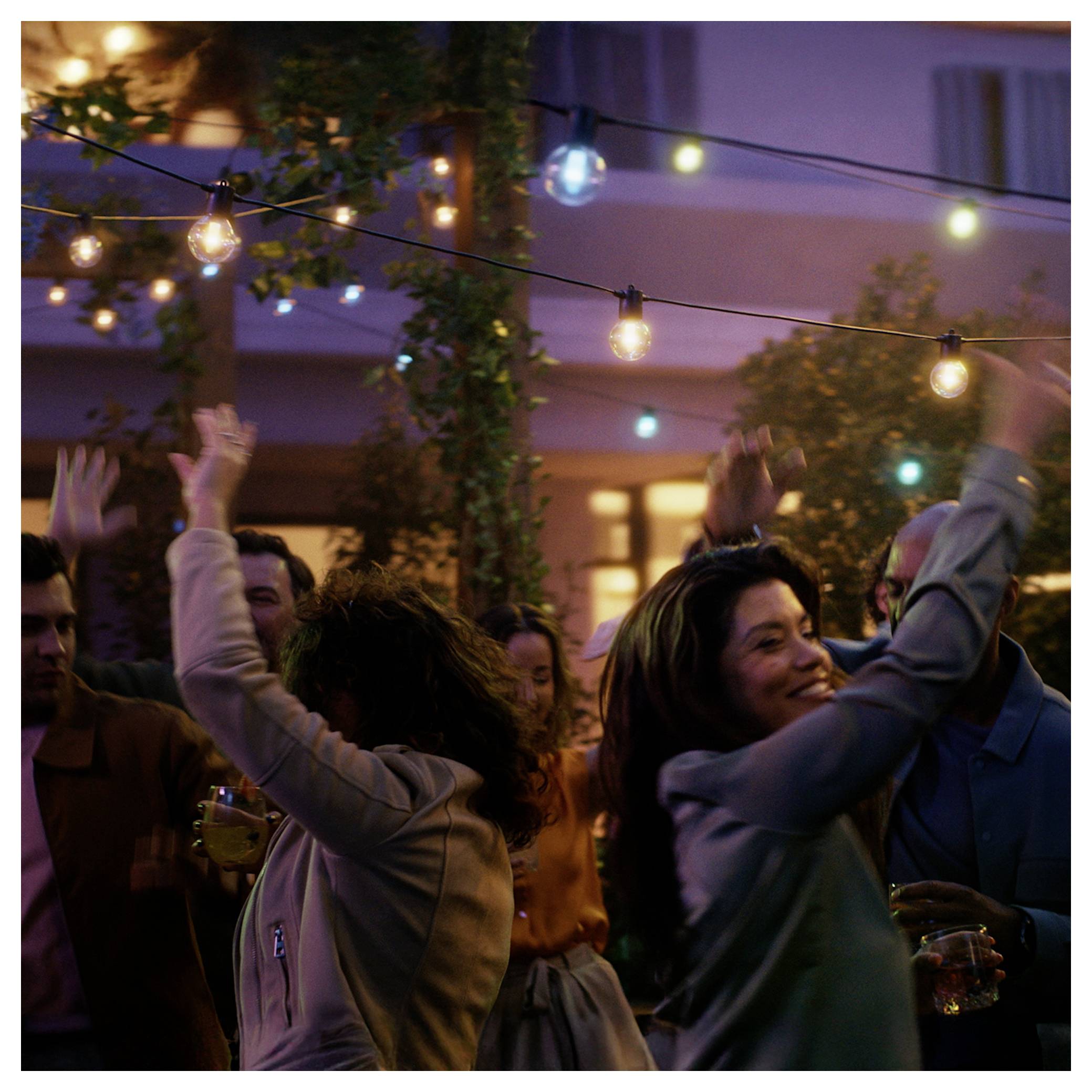 Des gens dansant joyeusement sous des guirlandes lumineuses lors d'une soirée en plein air, dans une ambiance festive, avec un bâtiment chaleureusement éclairé en arrière-plan.