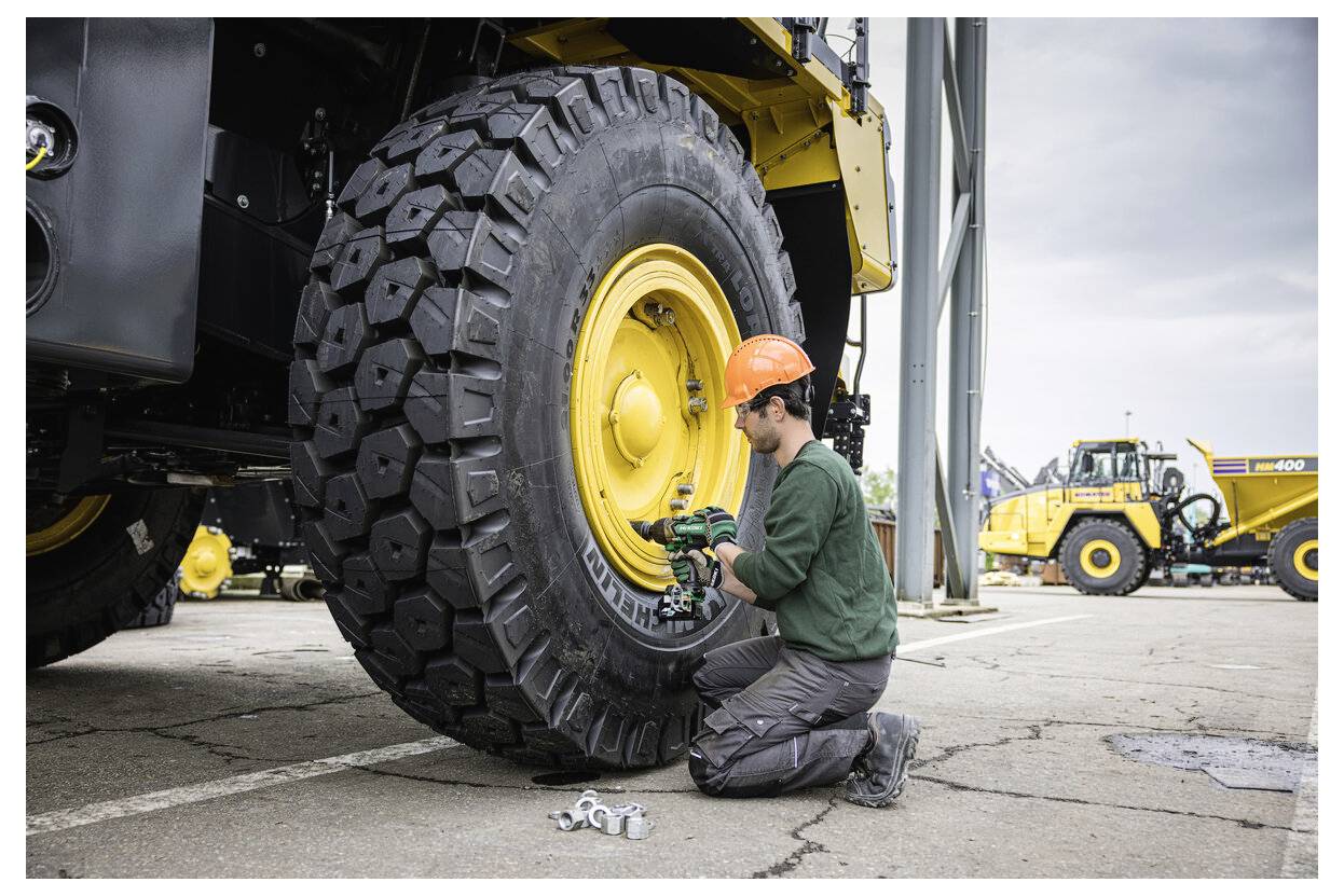 Une personne portant un casque orange répare un grand pneu jaune sur un engin de chantier. Un véhicule similaire est visible en arrière-plan.