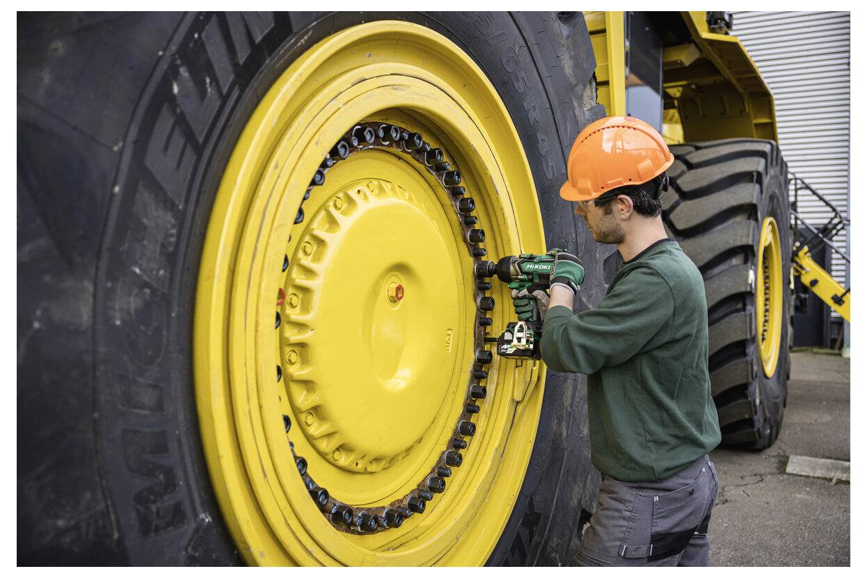 Un travailleur portant un casque de chantier resserre des boulons sur une grande roue de véhicule industriel jaune à l'aide d'un outil électrique.