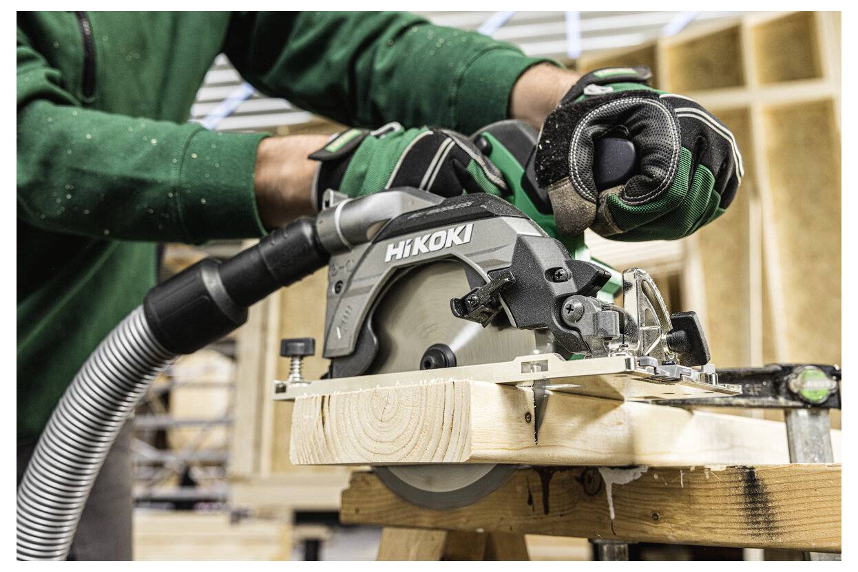 Personne portant une veste verte, utilisant une scie circulaire pour couper du bois, avec la sciure collectée par un tuyau d'aspirateur fixé dans un environnement d'atelier.