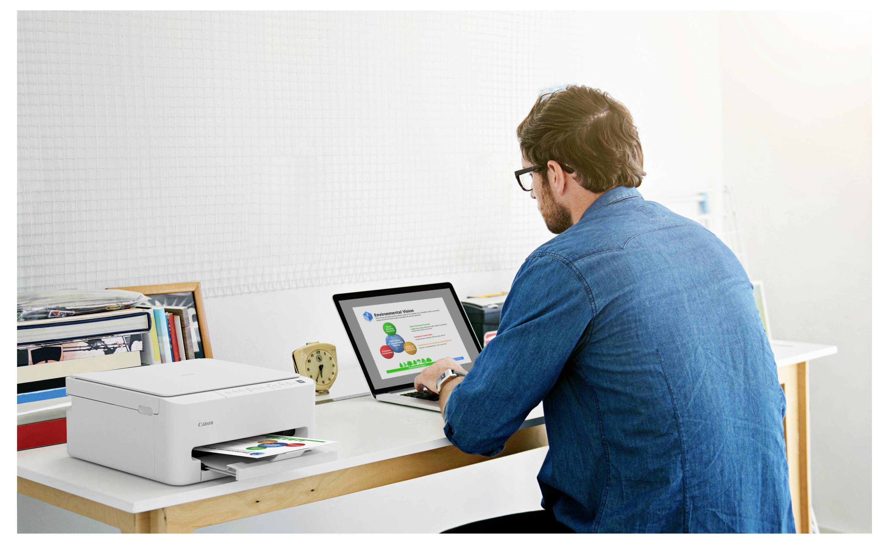 Une personne est assise à un bureau, utilisant un ordinateur portable avec une imprimante à côté d'elle. L'écran de l'ordinateur portable affiche un graphique ou une infographie coloré.