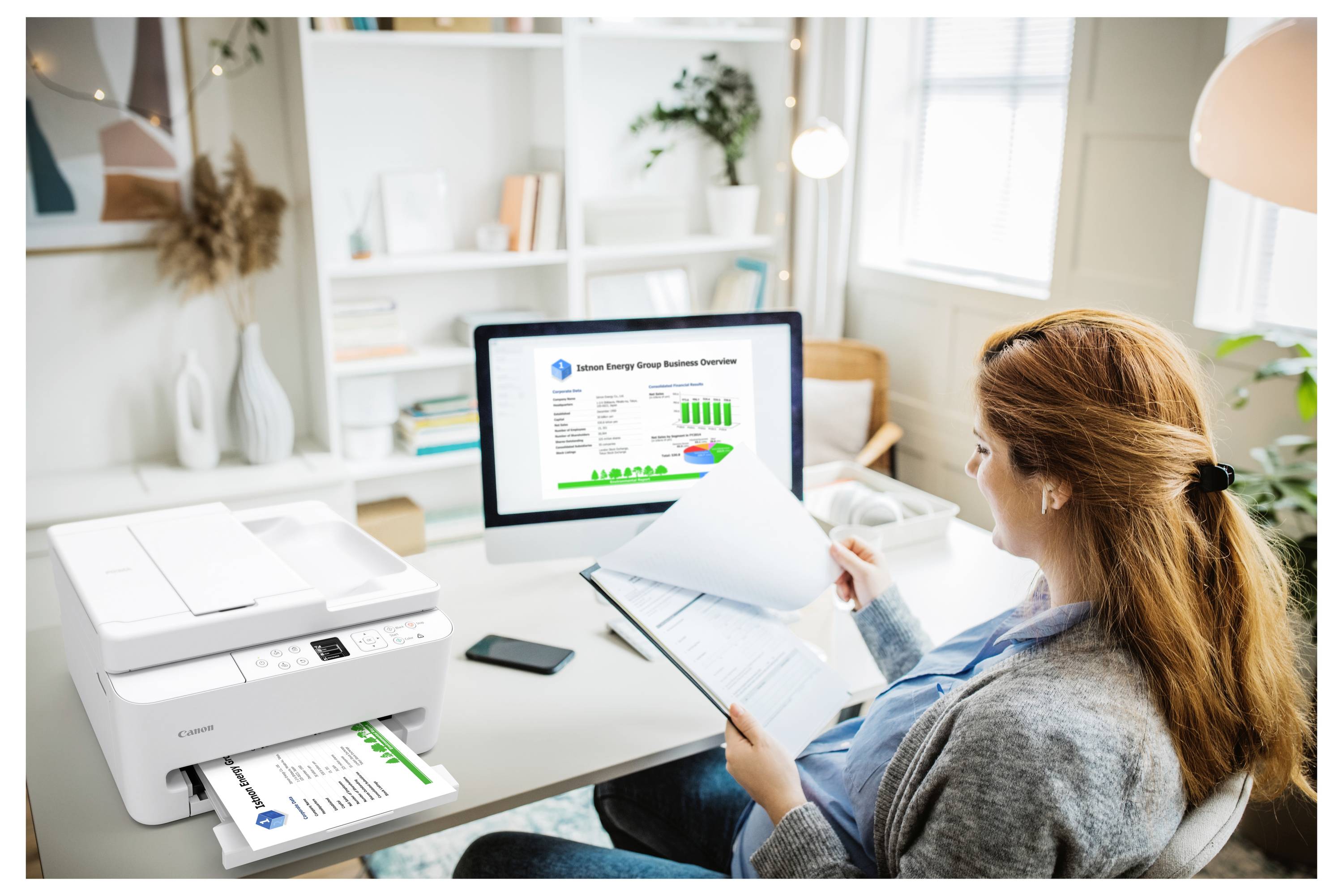 Une femme dans un bureau lit des documents. Un ordinateur de bureau affiche un graphique intitulé « Consommation énergétique annuelle » avec une imprimante à proximité qui distribue du papier.