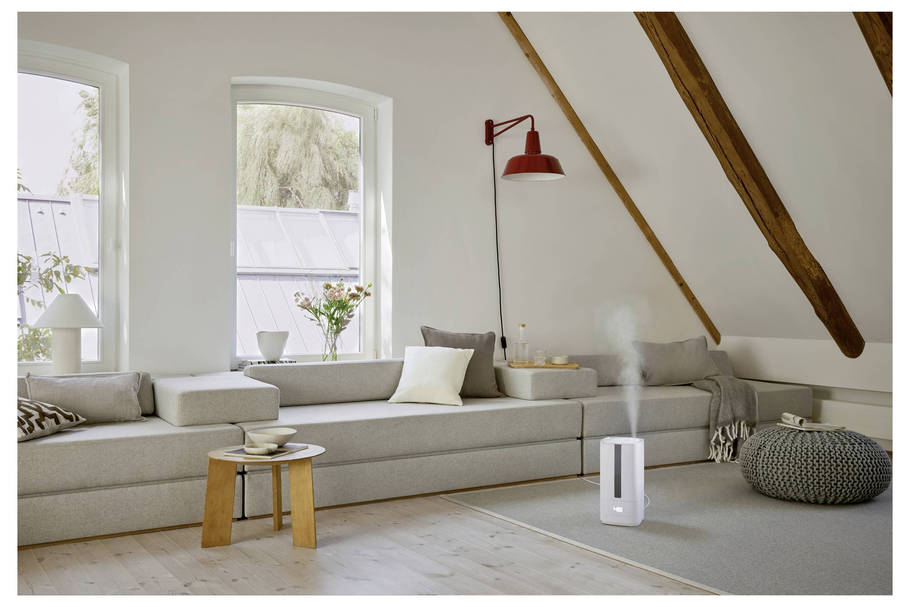 Un salon de combles douillet avec un long canapé gris, une lampe murale rouge, des coussins décoratifs et un humidificateur qui émet de la vapeur sur un tapis.