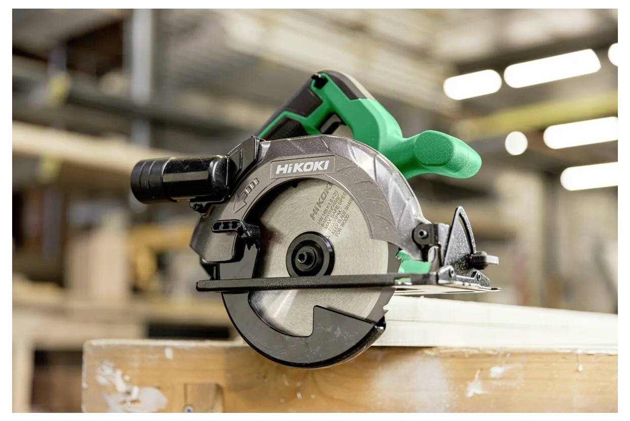 Une scie circulaire avec une poignée verte et la marque 'HiKOKI' sur le carter de lame repose sur une surface en bois dans un environnement d'atelier.