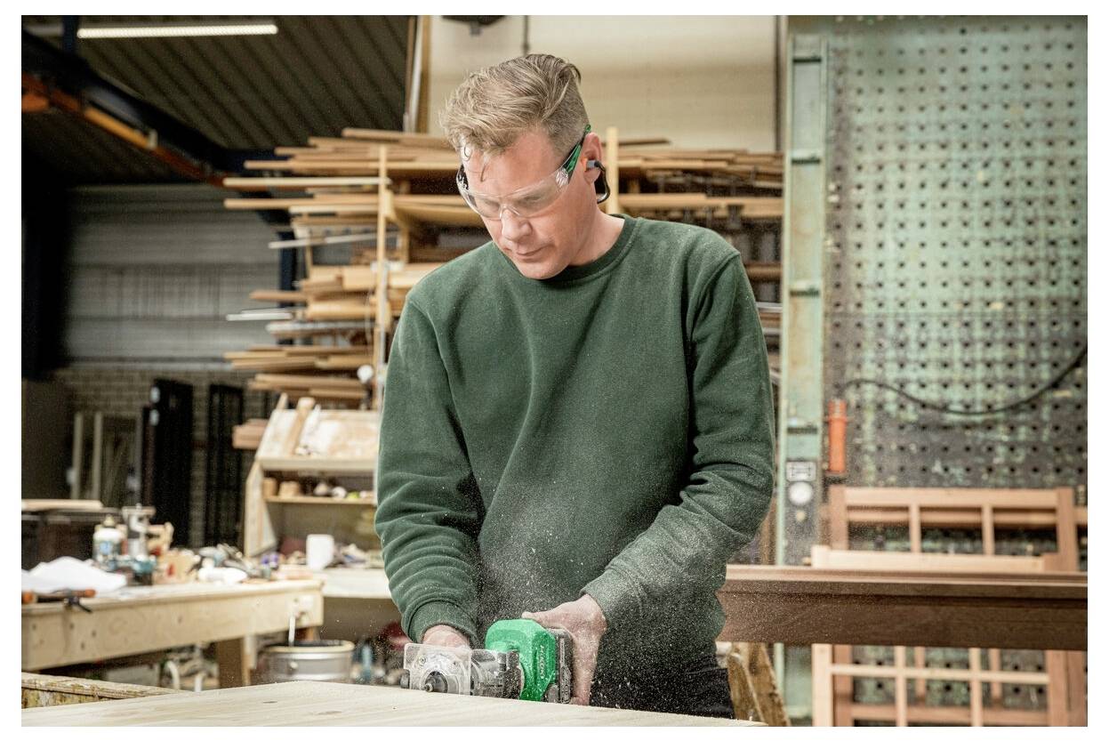 Une personne portant des lunettes de sécurité utilise un outil électrique dans un atelier de menuiserie, entourée de piles de bois et d'équipements d'atelier.