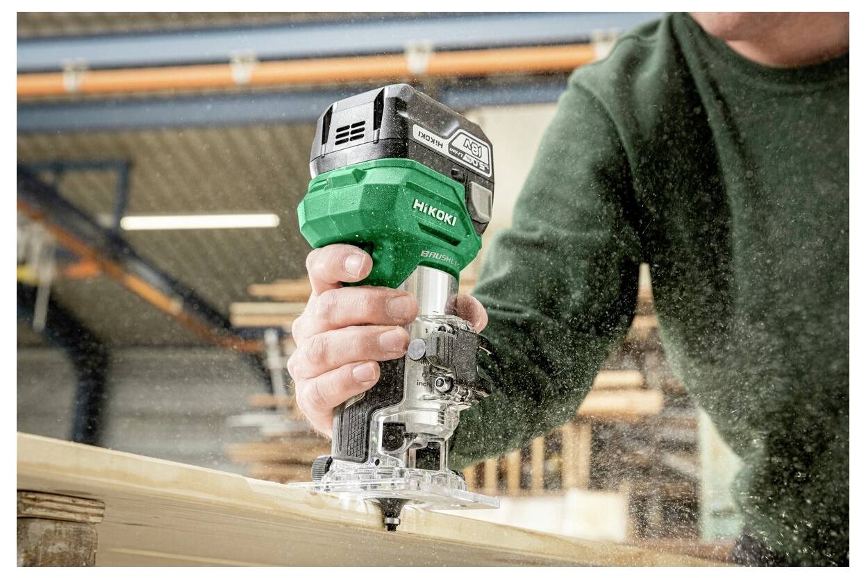 Une personne utilise un routeur électrique à main vert et noir sur une surface en bois dans un environnement d'atelier, provoquant la dispersion de poussière de bois.