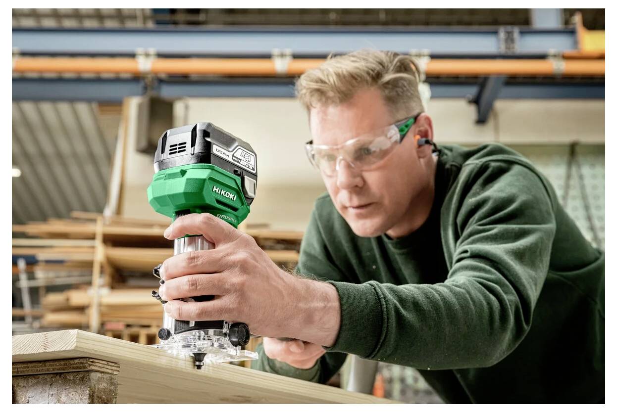 Une personne portant des lunettes de sécurité utilise une défonceuse manuelle verte et noire sur une planche de bois dans un environnement d'atelier.