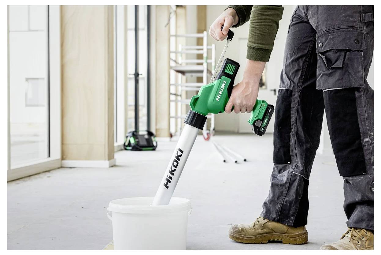 Une personne utilise un outil électrique sans fil vert et blanc pour mélanger du contenu dans un seau à l'intérieur d'une pièce en cours de construction partiellement achevée.
