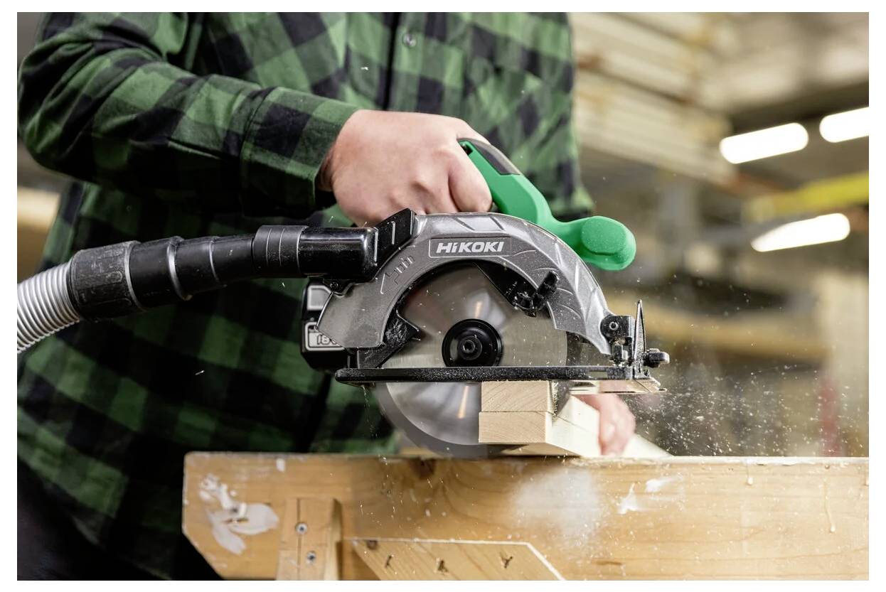 Une personne portant une chemise à carreaux verts utilise une scie circulaire pour couper un morceau de bois sur un établi, avec des copeaux de bois en mouvement.
