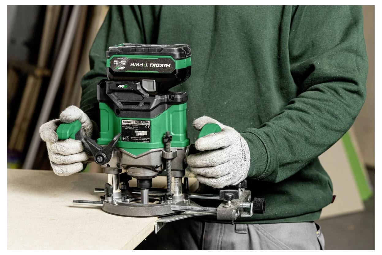Une personne portant des gants utilise un routeur vert sur une planche en bois, concentrée sur l'artisanat ou le travail du bois dans un environnement d'atelier.