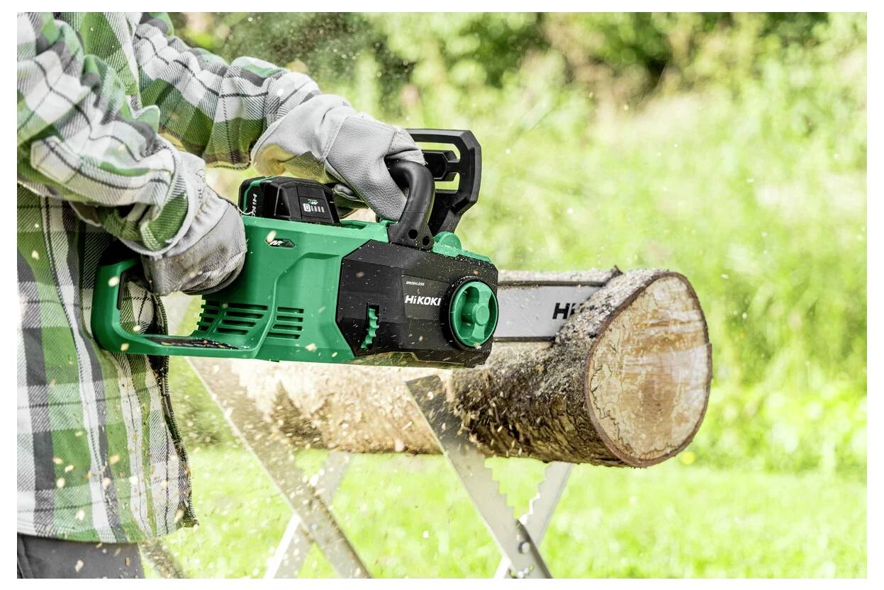 Une personne portant des gants utilise une tronçonneuse verte pour couper un journal sur un chevalet à l'extérieur, avec des copeaux de bois volant autour.