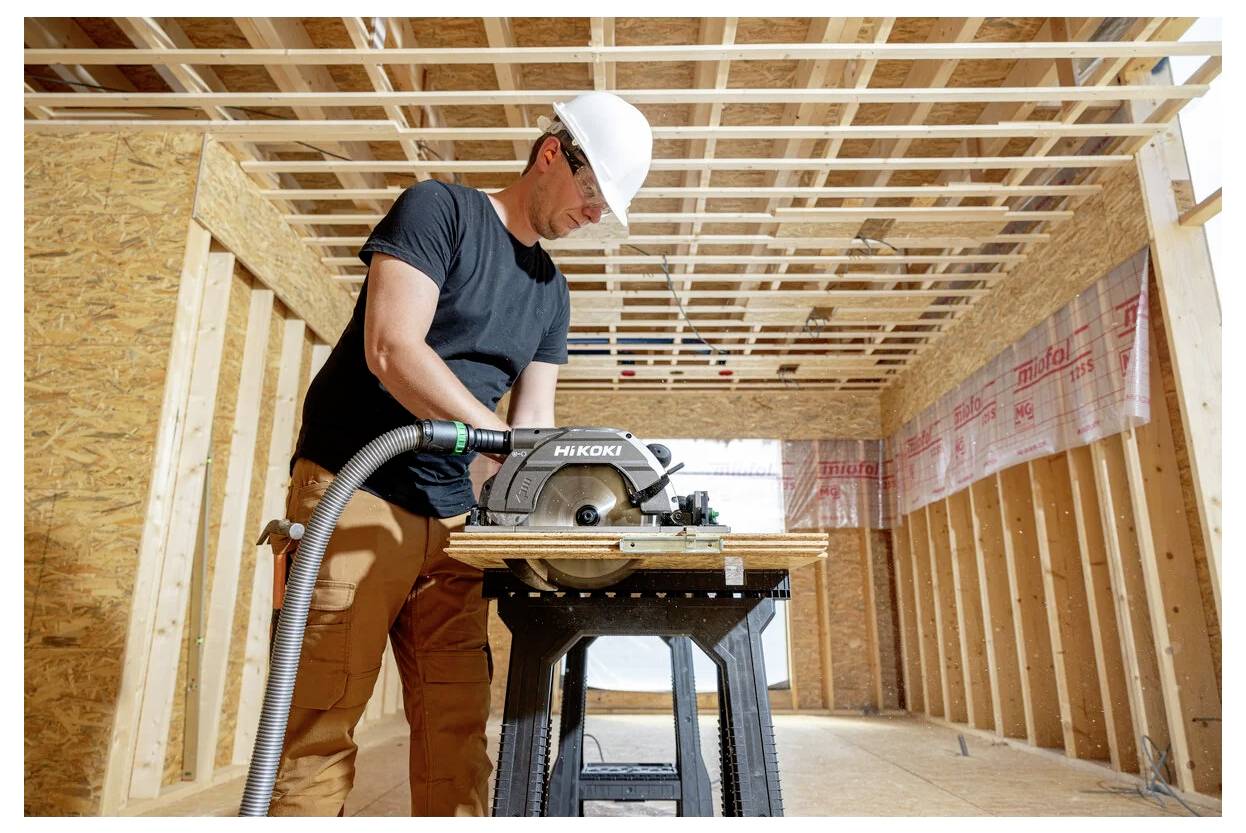 Un ouvrier du bâtiment portant un casque utilise une scie circulaire à l'intérieur d'une structure en bois partiellement construite, concentré sur sa tâche.