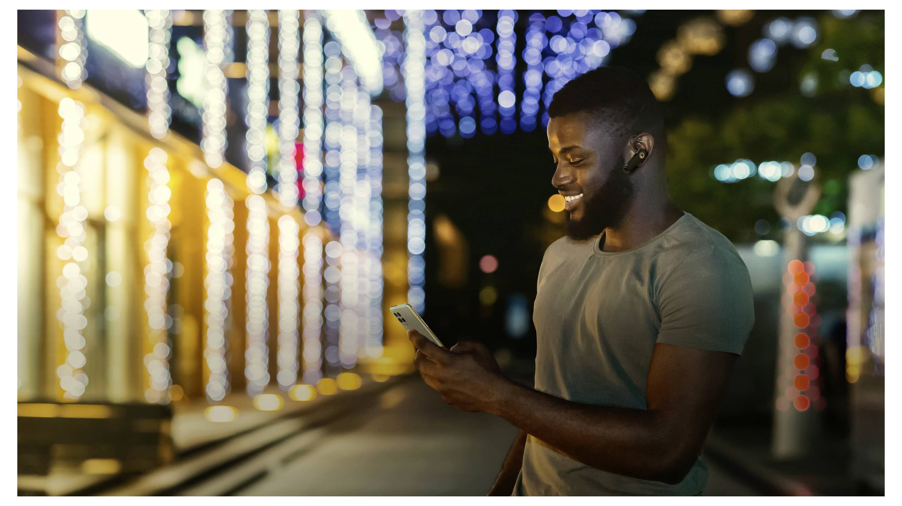 Un homme souriant, regardant son téléphone, debout dehors la nuit, avec des lumières festives ornant les bâtiments en arrière-plan.