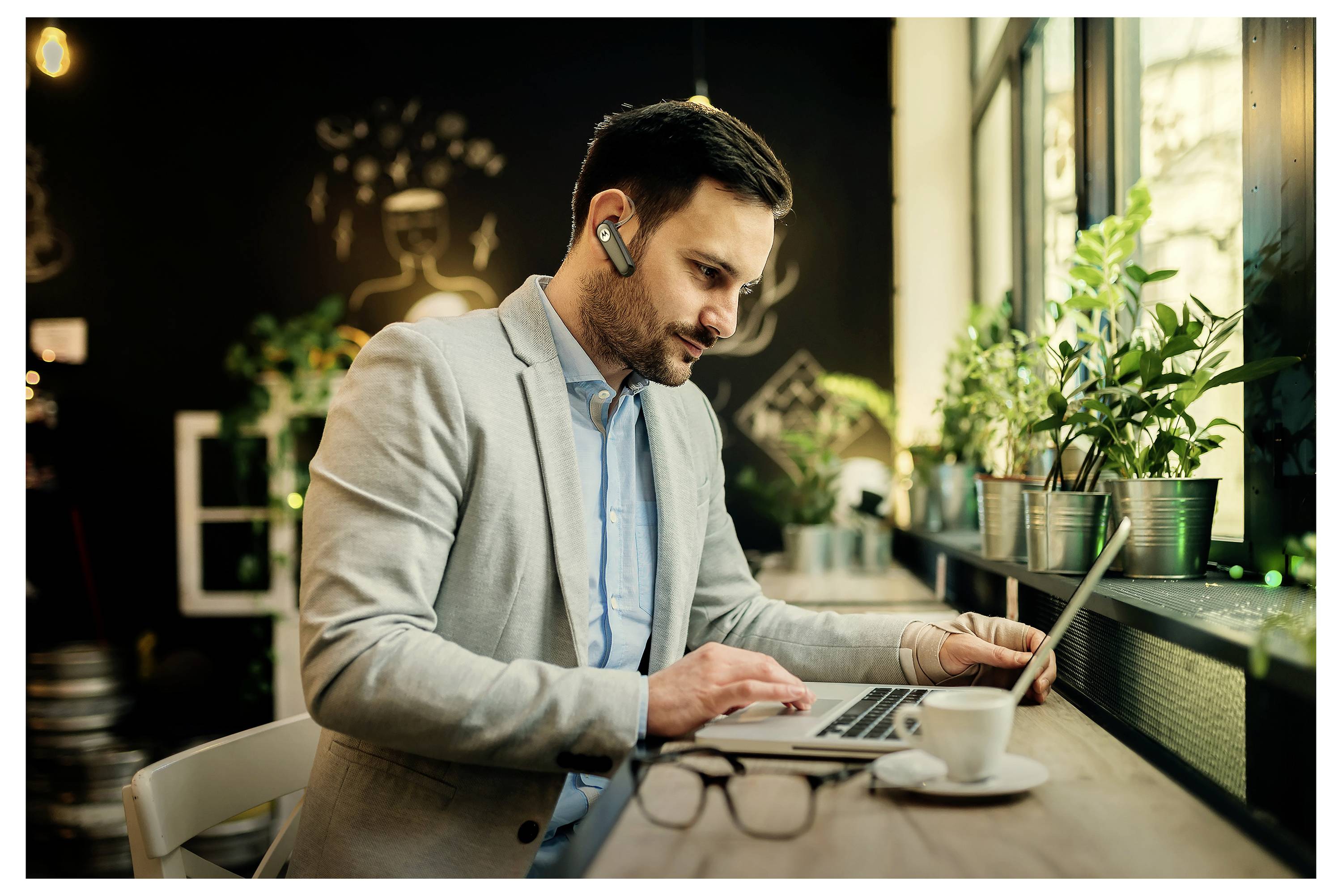 Un homme portant un blazer gris travaille sur un ordinateur portable dans un café accueillant. Il porte un écouteur et est assis près d'une fenêtre, entouré de plantes.