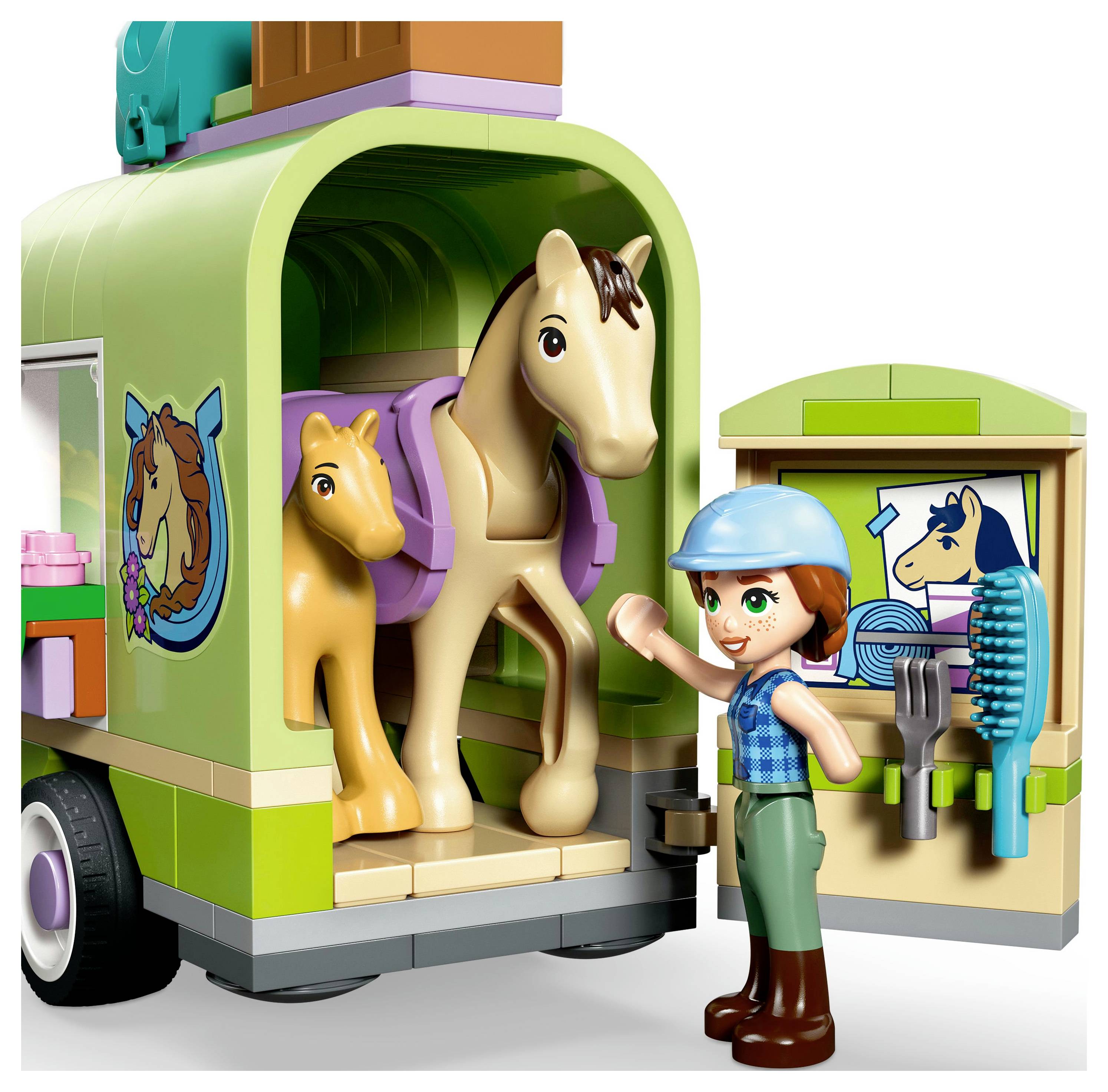 Un enfant portant un casque fait un signe de la main joyeusement à côté d'une remorque pour chevaux miniatures contenant deux chevaux de jouet, avec des outils de toilettage disposés sur une porte ouverte.