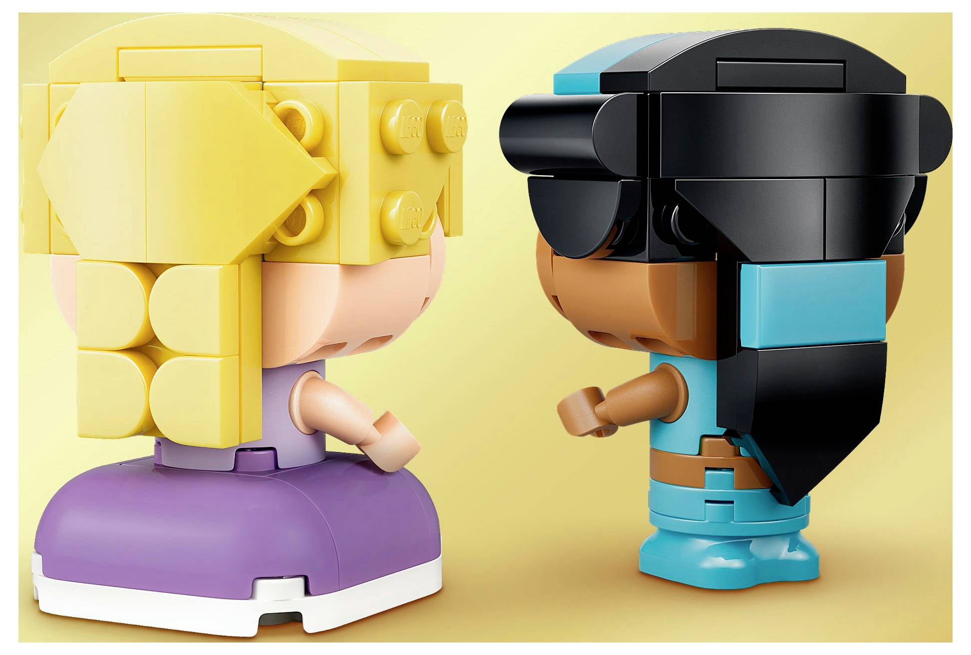 Deux figurines de jouet se font face sur un fond jaune. La figurine de gauche a une tête jaune et une base violette. La figurine de droite a une base bleue et de longs cheveux noirs.