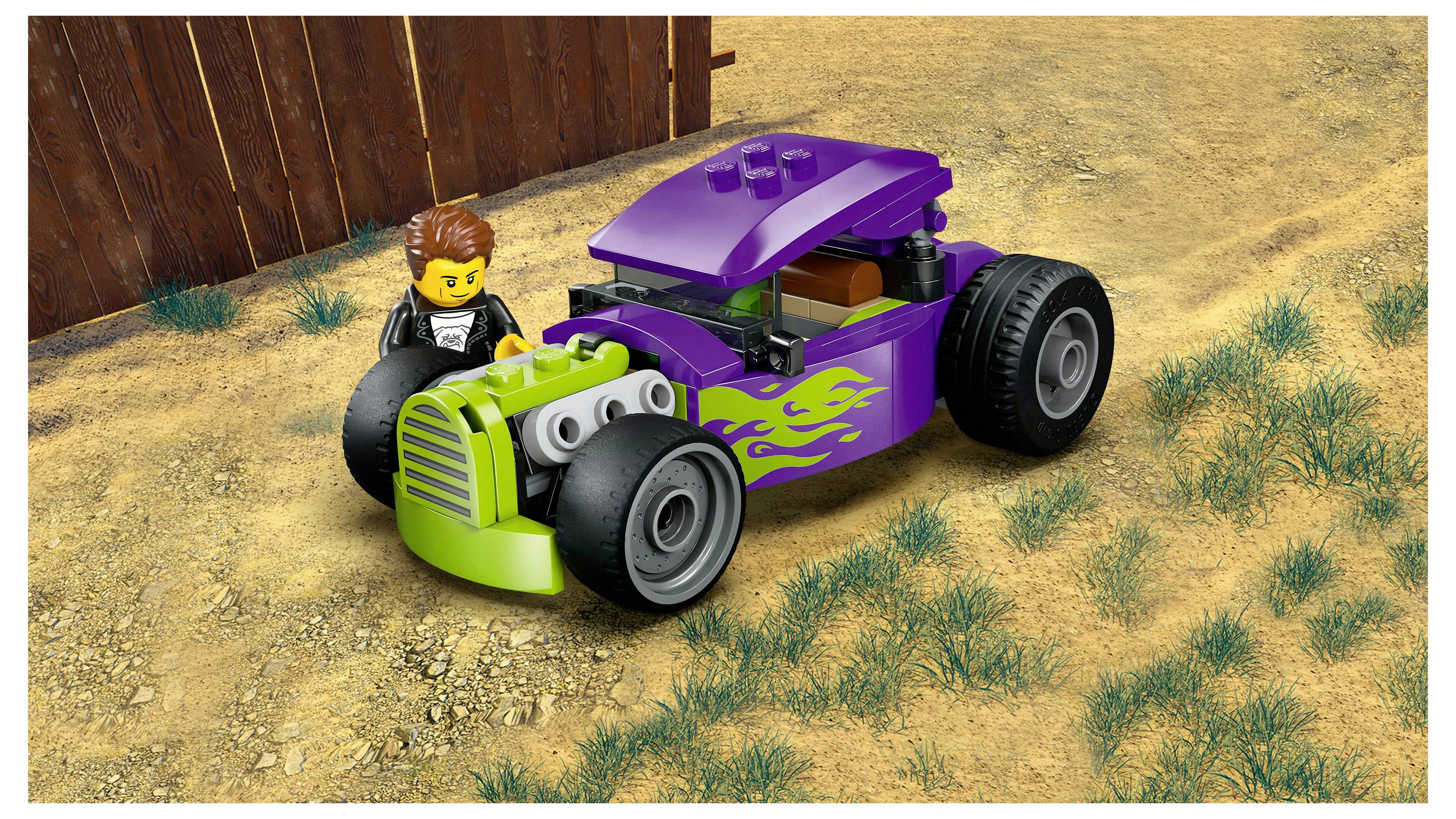 Une figurine de jouet est debout à côté d'un hot-rod LEGO violet et vert avec des décalcomanies de flammes sur une surface terreuse, près d'une clôture en bois.