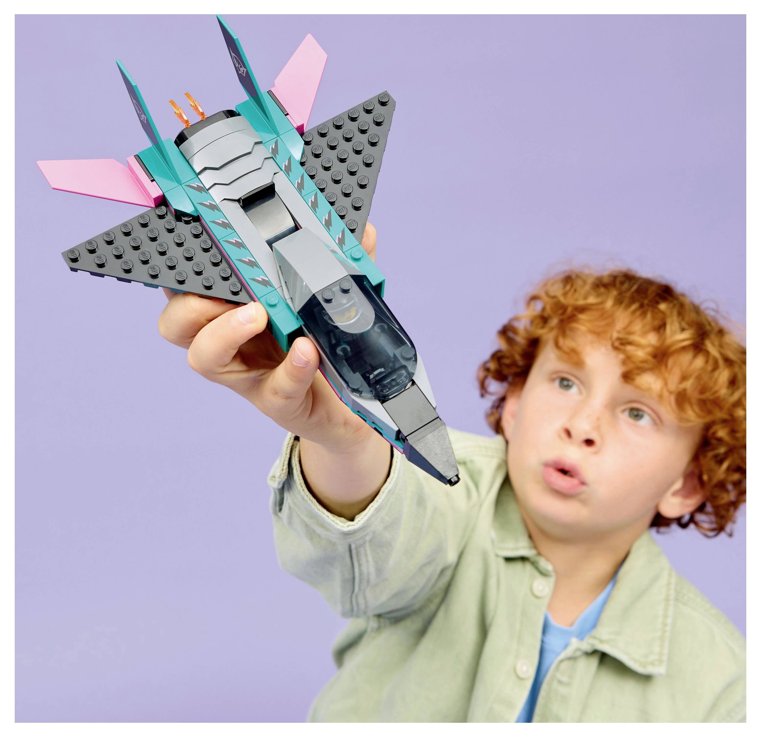 Un enfant tient avec enthousiasme une fusée jouet, conçue avec des couleurs vives et des ailes élégantes, devant un fond mauve pastel.