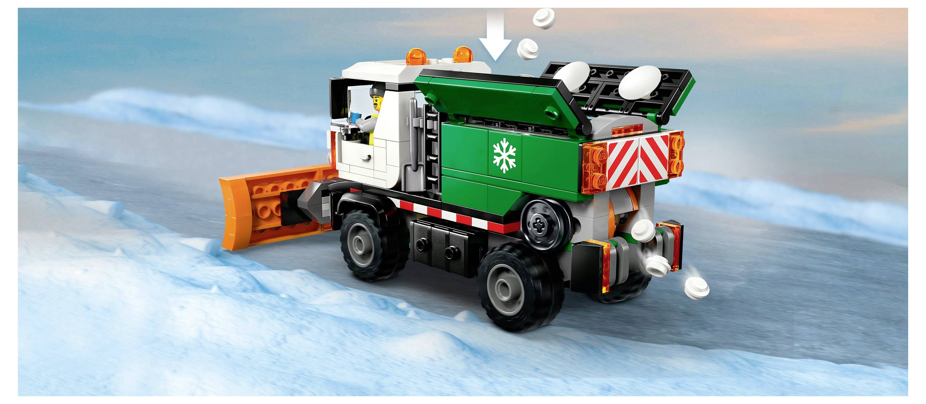 Un camion chasse-neige jouet sur une route enneigée distribue des boules de neige. Le camion est vert et blanc avec une lame chasse-neige orange à l'avant.