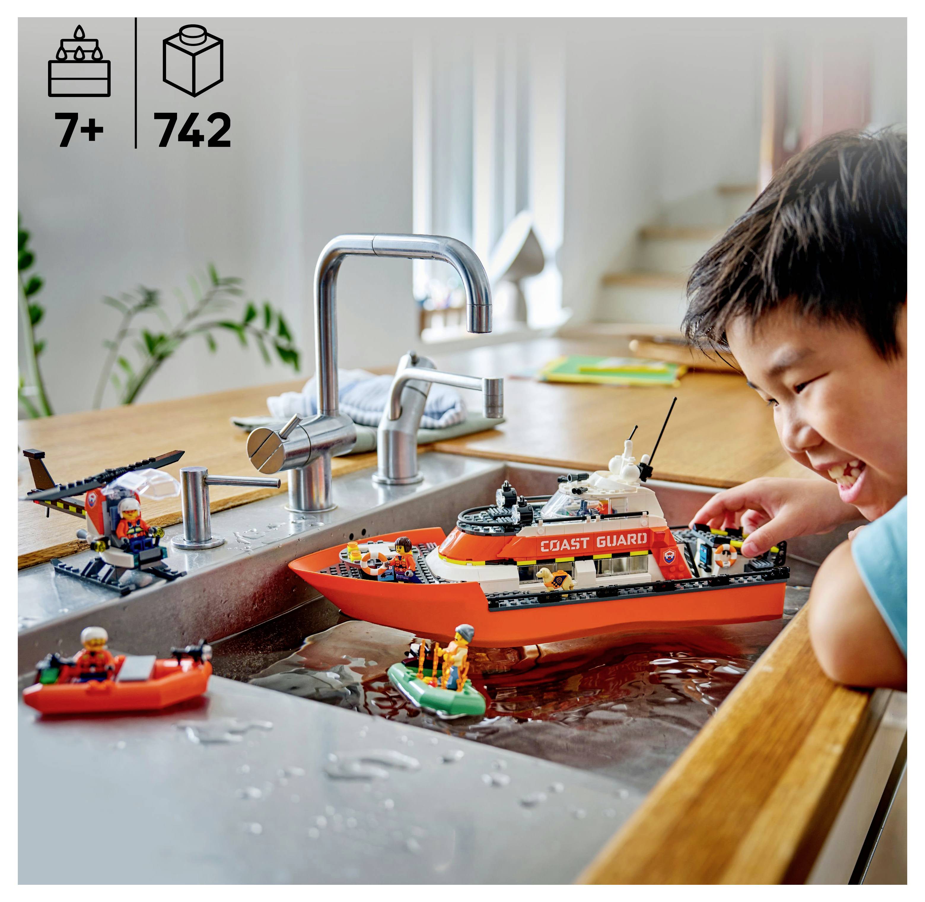 Un enfant joue joyeusement avec un ensemble LEGO de la Garde côtière dans un évier de cuisine rempli d'eau. L'ensemble comprend un bateau et un hélicoptère.