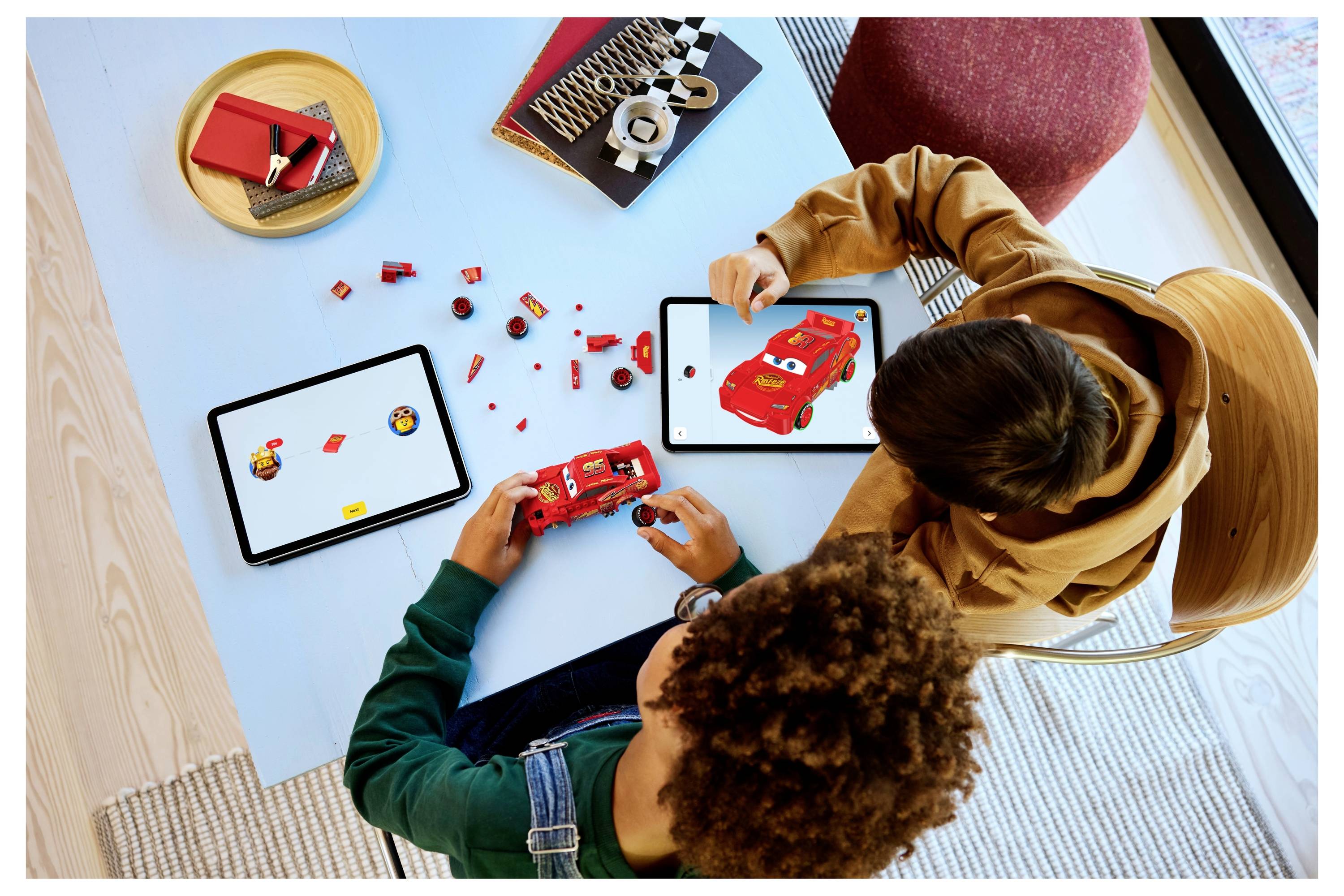 Deux enfants sont assis à une table en train d'assembler des voitures LEGO avec des briques rouges, en utilisant des tablettes affichant des instructions connexes.