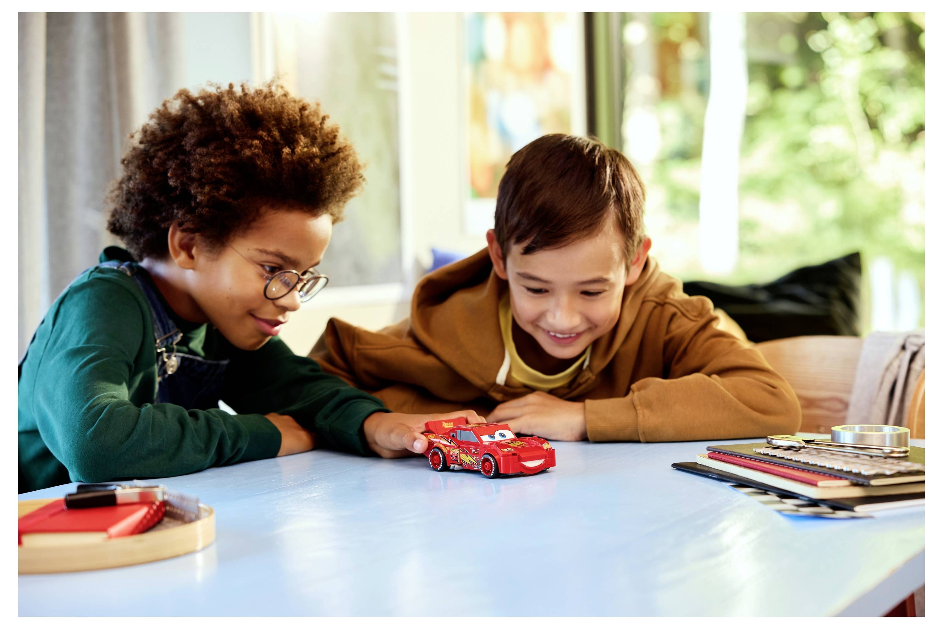 Deux enfants jouant joyeusement avec une voiture rouge sur une table, entourés de matériel artistique, dans une pièce lumineuse avec des arbres verts visibles à l'extérieur.