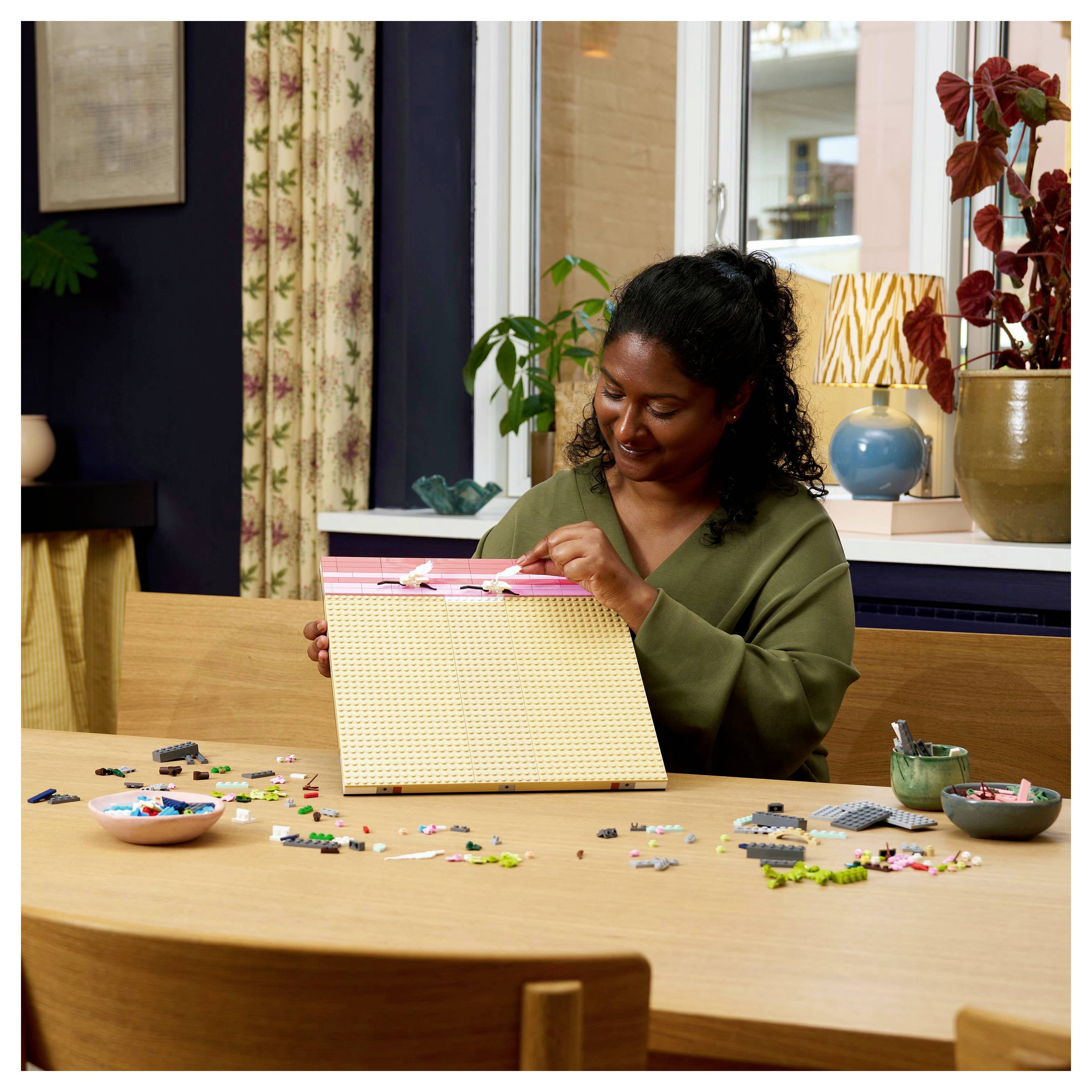 Une personne est assise à une table en train d'assembler une œuvre d'art en blocs colorés, entourée de blocs éparpillés et d'une plante en arrière-plan.