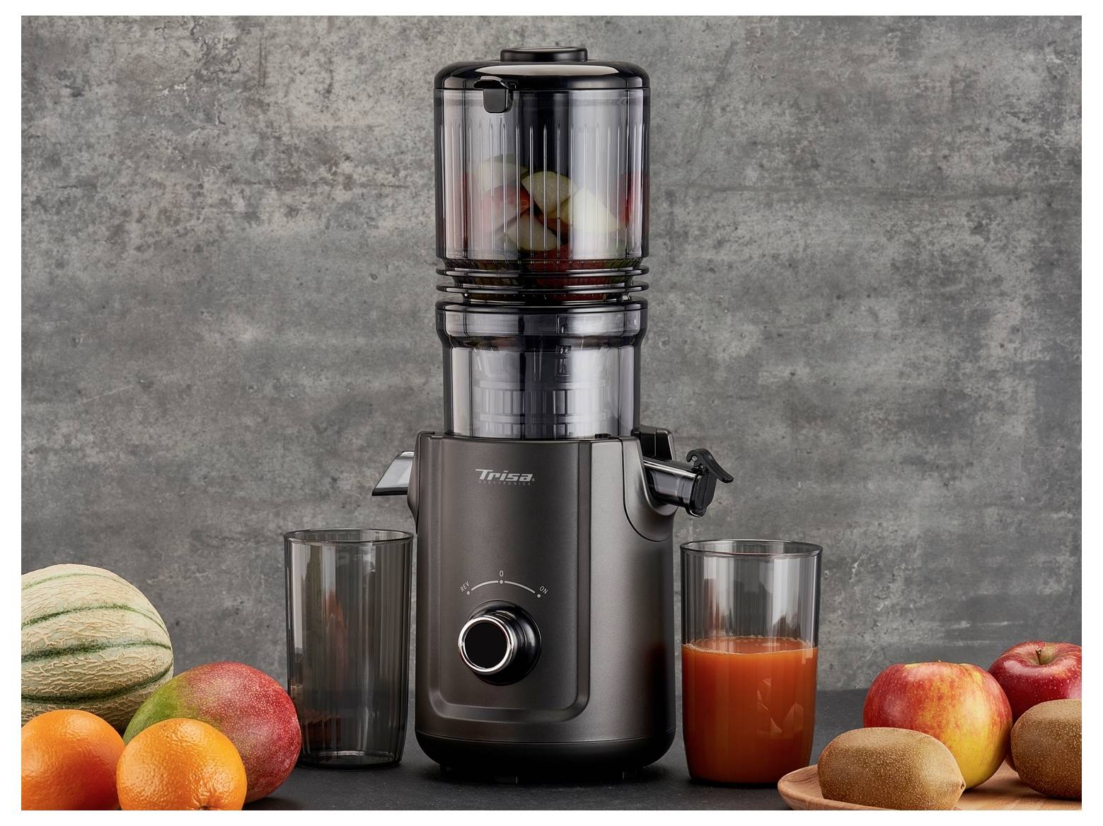 Un presse-fruits noir contenant des fruits à l'intérieur, entouré d'une variété de fruits frais et d'un verre de jus, sur un fond gris.