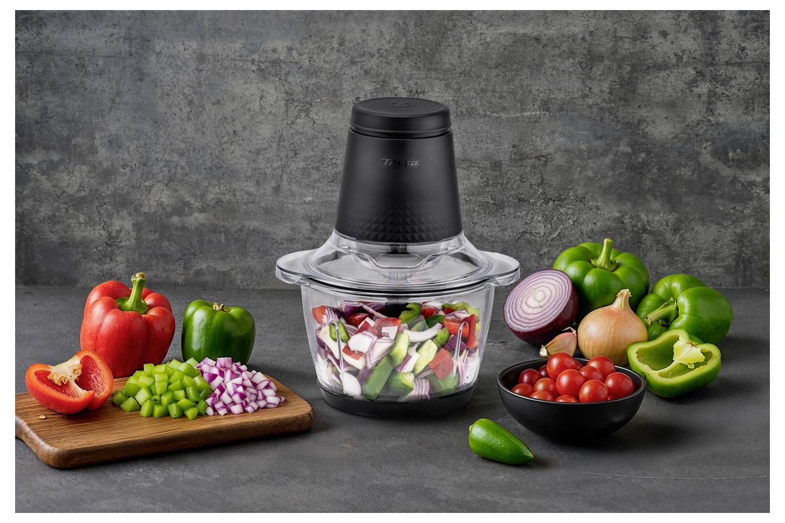 Un robot culinaire entouré de légumes coupés, comprenant des poivrons rouges et verts, un oignon rouge et des tomates cerises, sur un plan de travail.