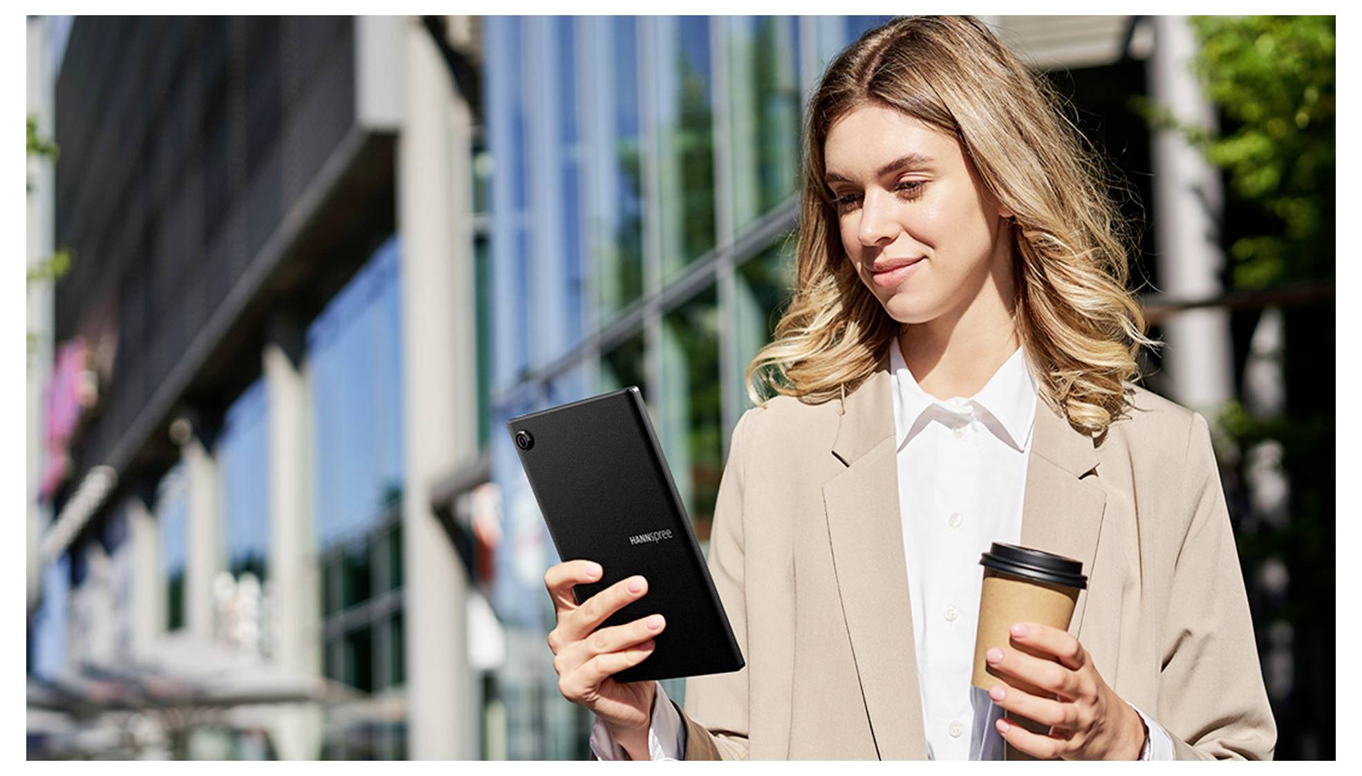 Une femme se tient dehors devant un bâtiment moderne, tenant une tablette et une tasse de café, souriante et regardant l'écran.