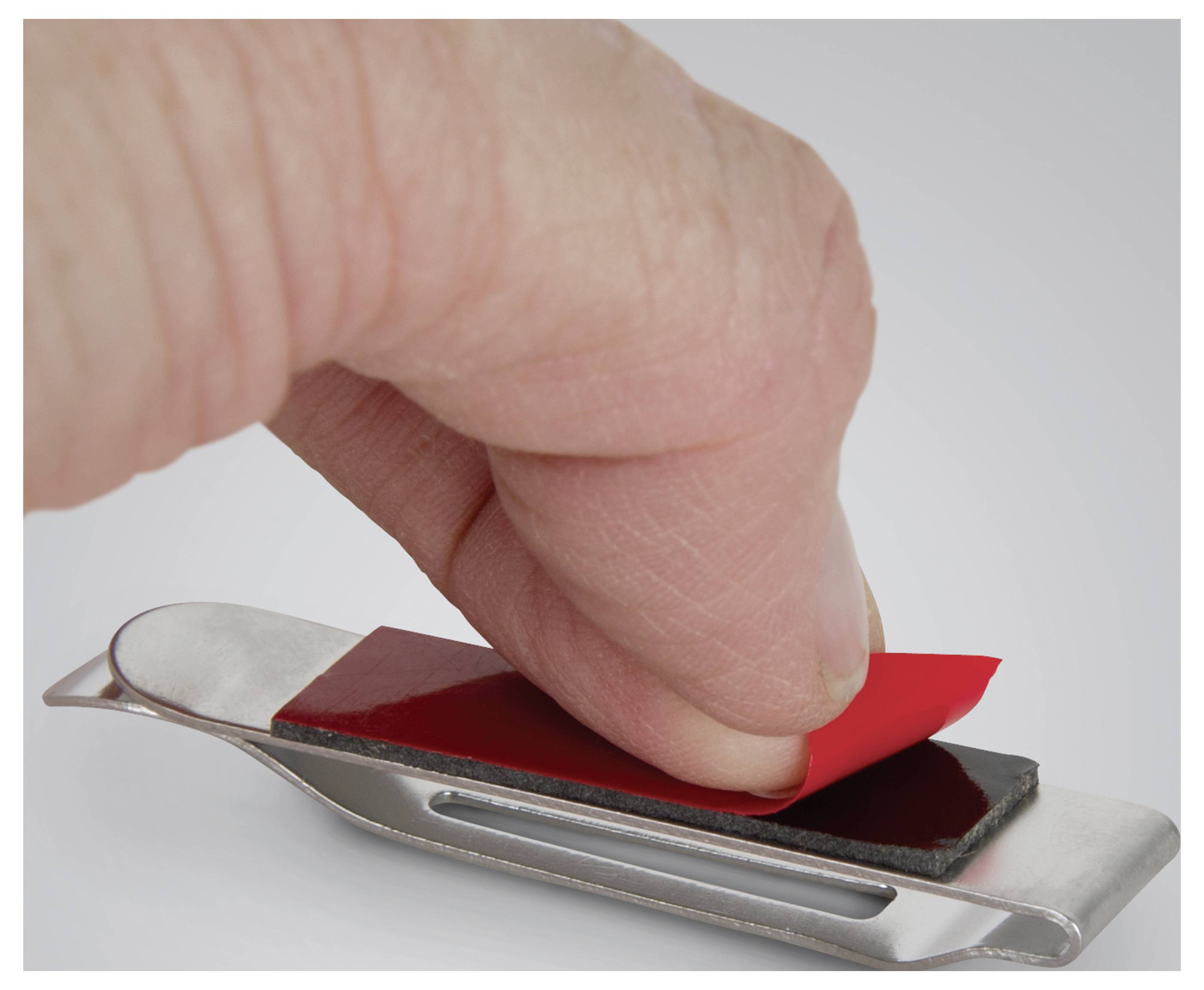 Une main retire un ruban adhésif rouge d'un petit outil métallique plat, qui semble être un grattoir ou un décolleur d'étiquettes, sur un fond gris.