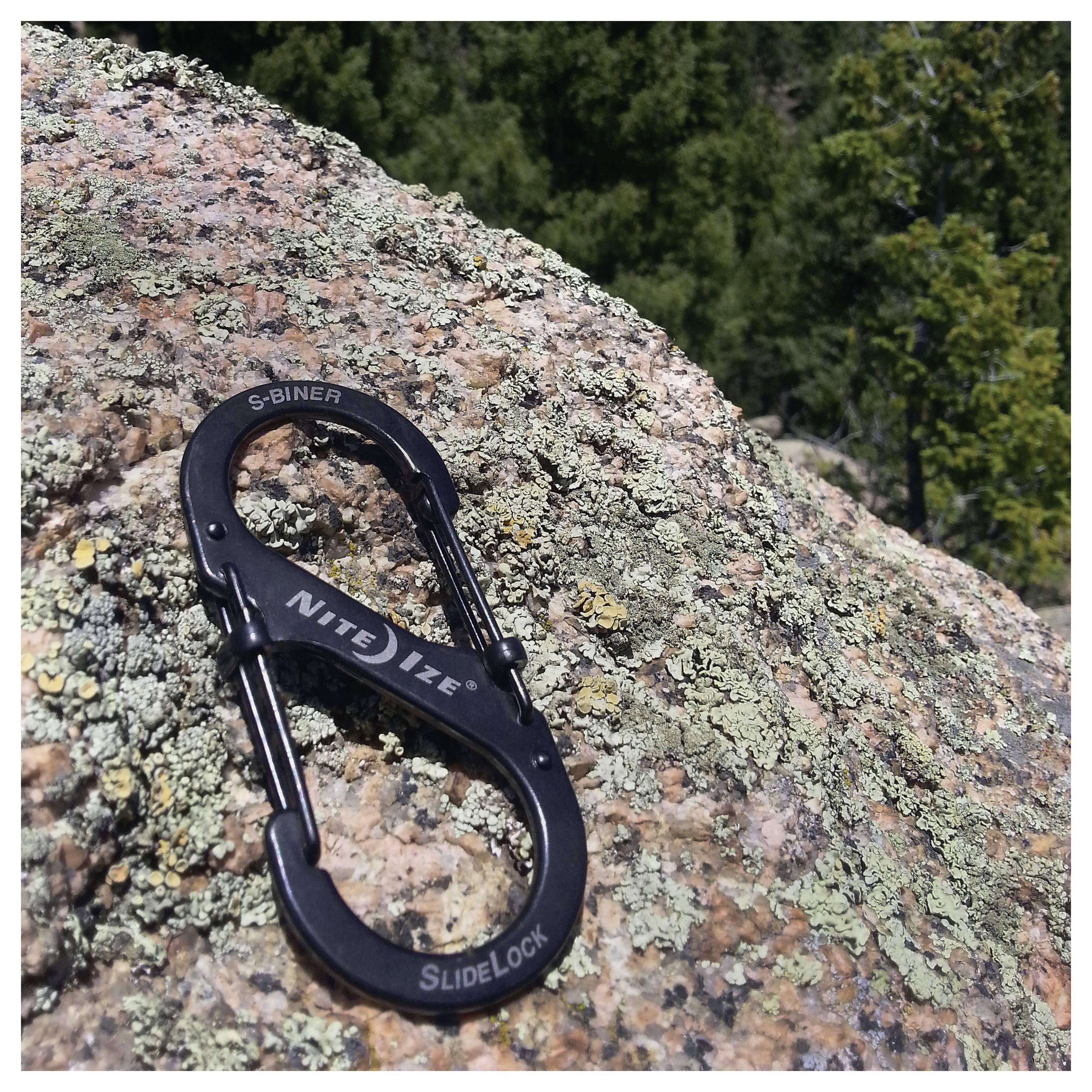 Un mousqueton noir 'Nite Ize S-Biner SlideLock' repose sur un rocher couvert de lichen, sur fond naturel flou.