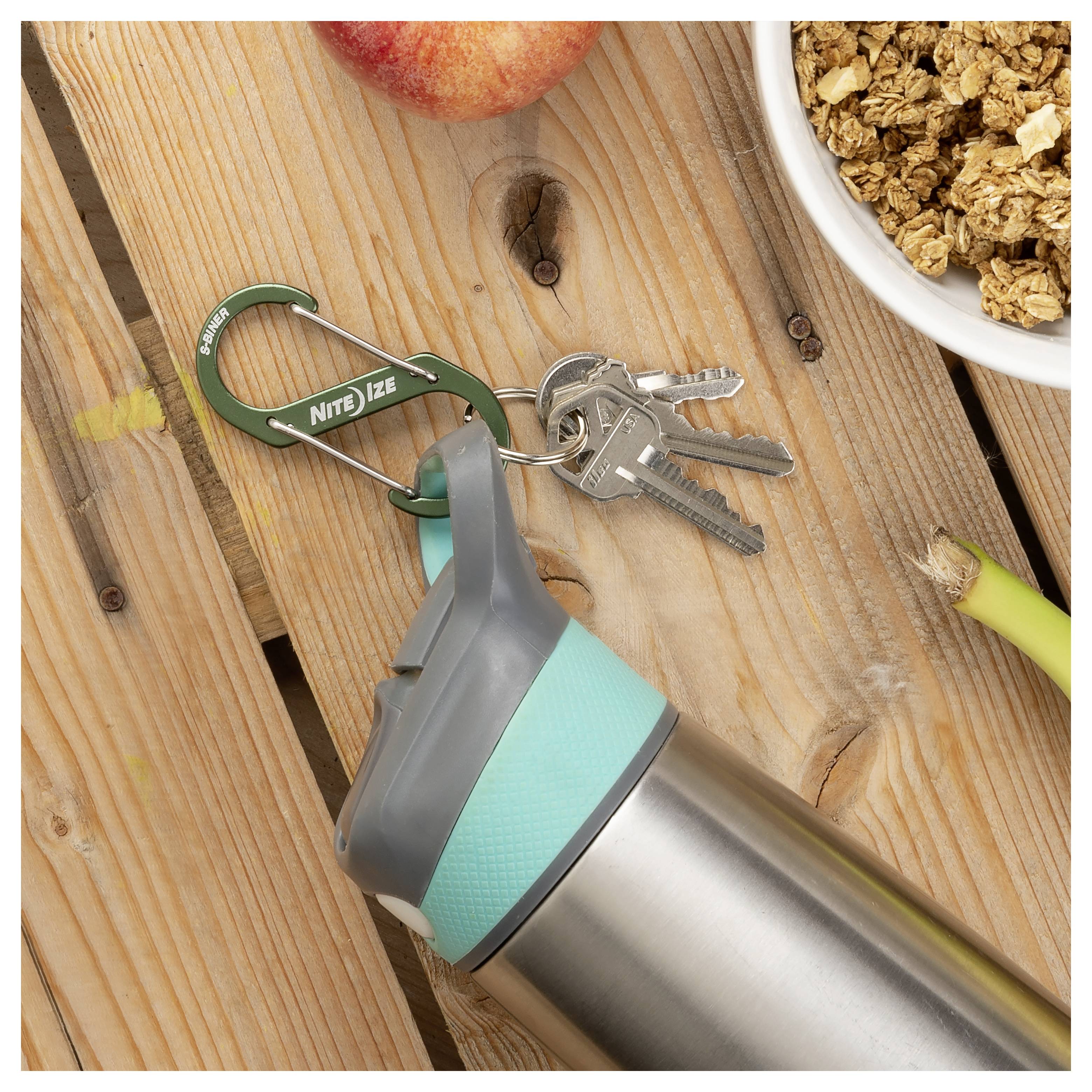 Une bouteille d'eau réutilisable avec un couvercle bleu-vert est posée sur une table en bois à côté d'un trousseau de clés attaché à un mousqueton vert, une pomme et un bol de granola.