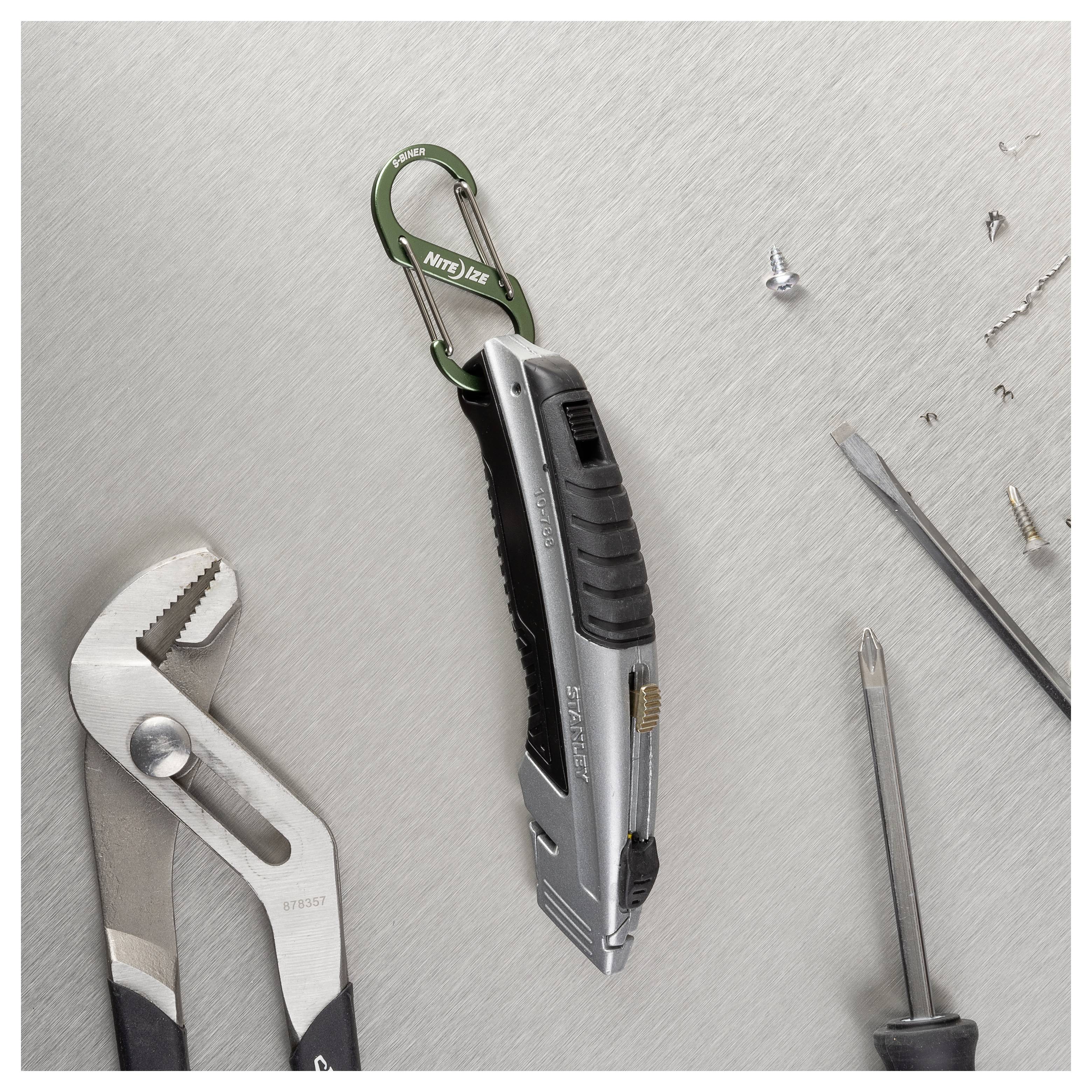 Couteau utilitaire sur une surface métallique, attaché à un mousqueton vert. Autour se trouvent des pinces, un tournevis et des vis desserrées.