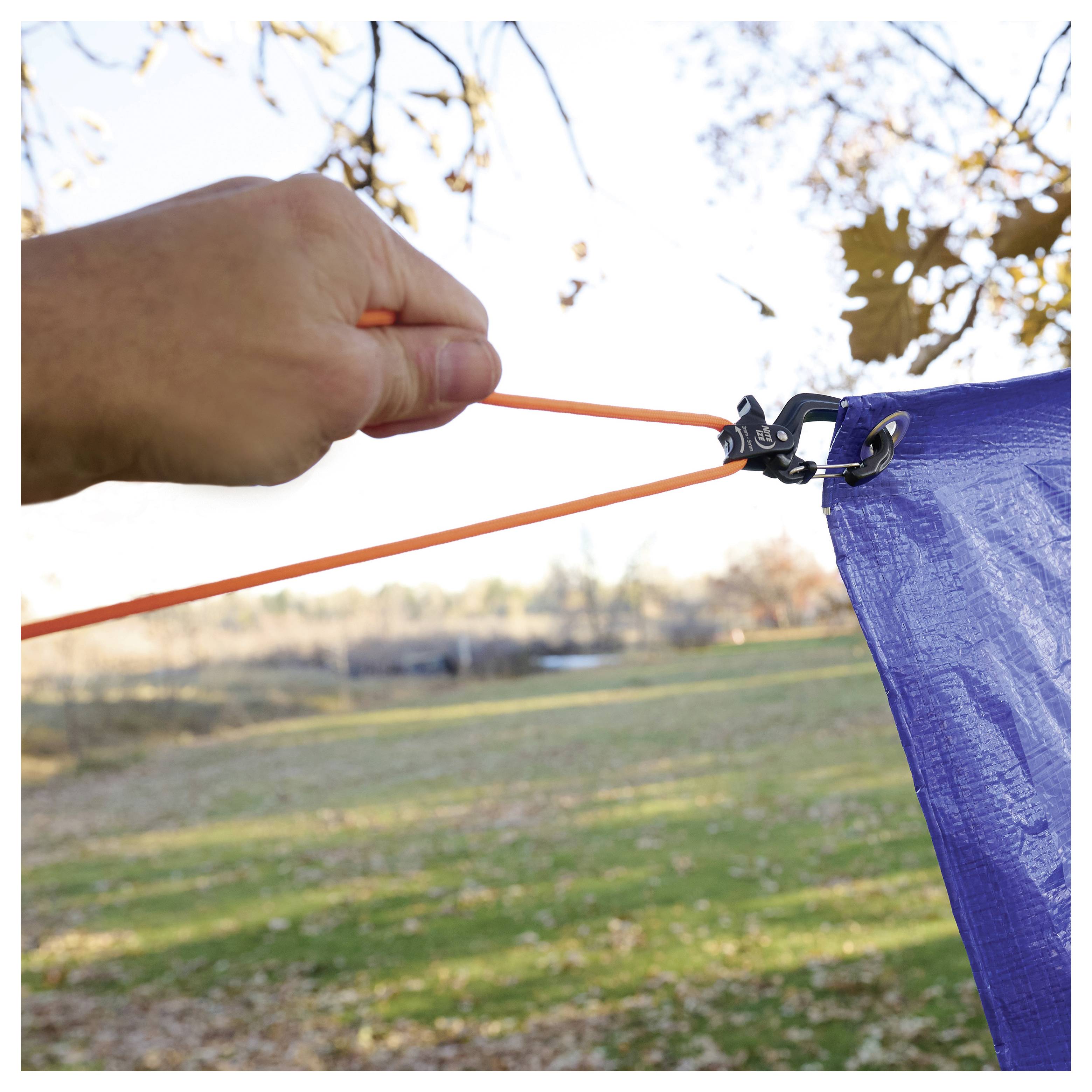 Une main est visible en train de tirer un cordon orange pour tendre une bâche bleue, la fixant sur un attelage, avec un paysage d'automne en arrière-plan.