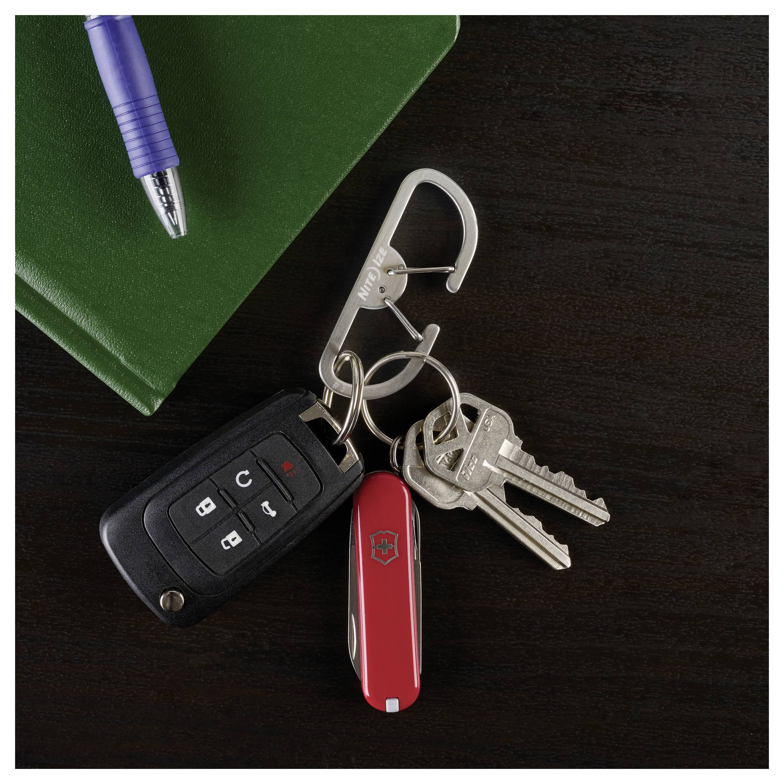 Un trousseau avec un couteau de poche rouge et un porte-clés de voiture sont attachés à un mousqueton argenté. Un carnet vert et un stylo bleu sont posés à proximité sur une surface sombre.