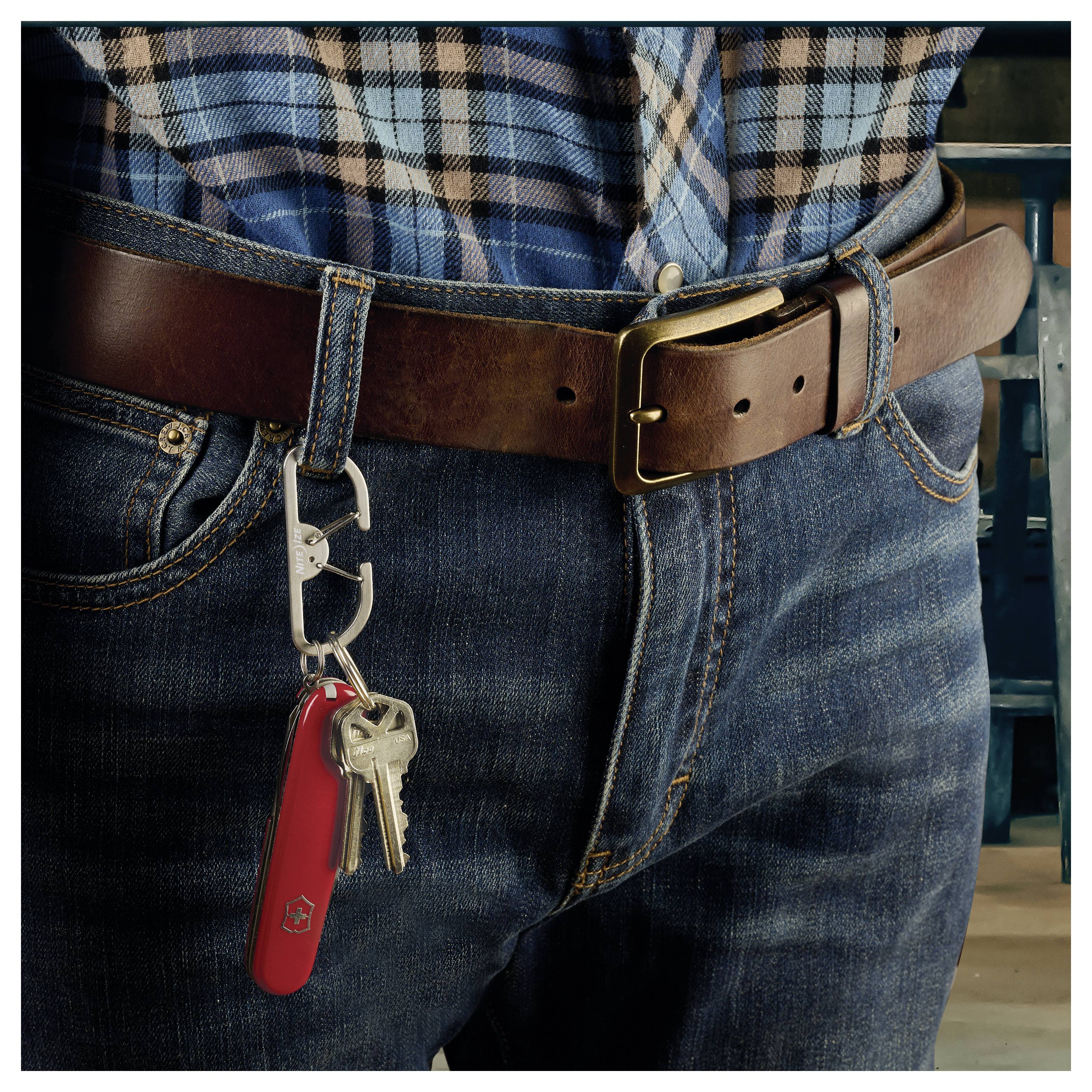 Une personne portant un jean et une chemise à carreaux, avec une ceinture en cuir sur laquelle sont accrochés des clés et un couteau de poche rouge fixé à une passant de pantalon.