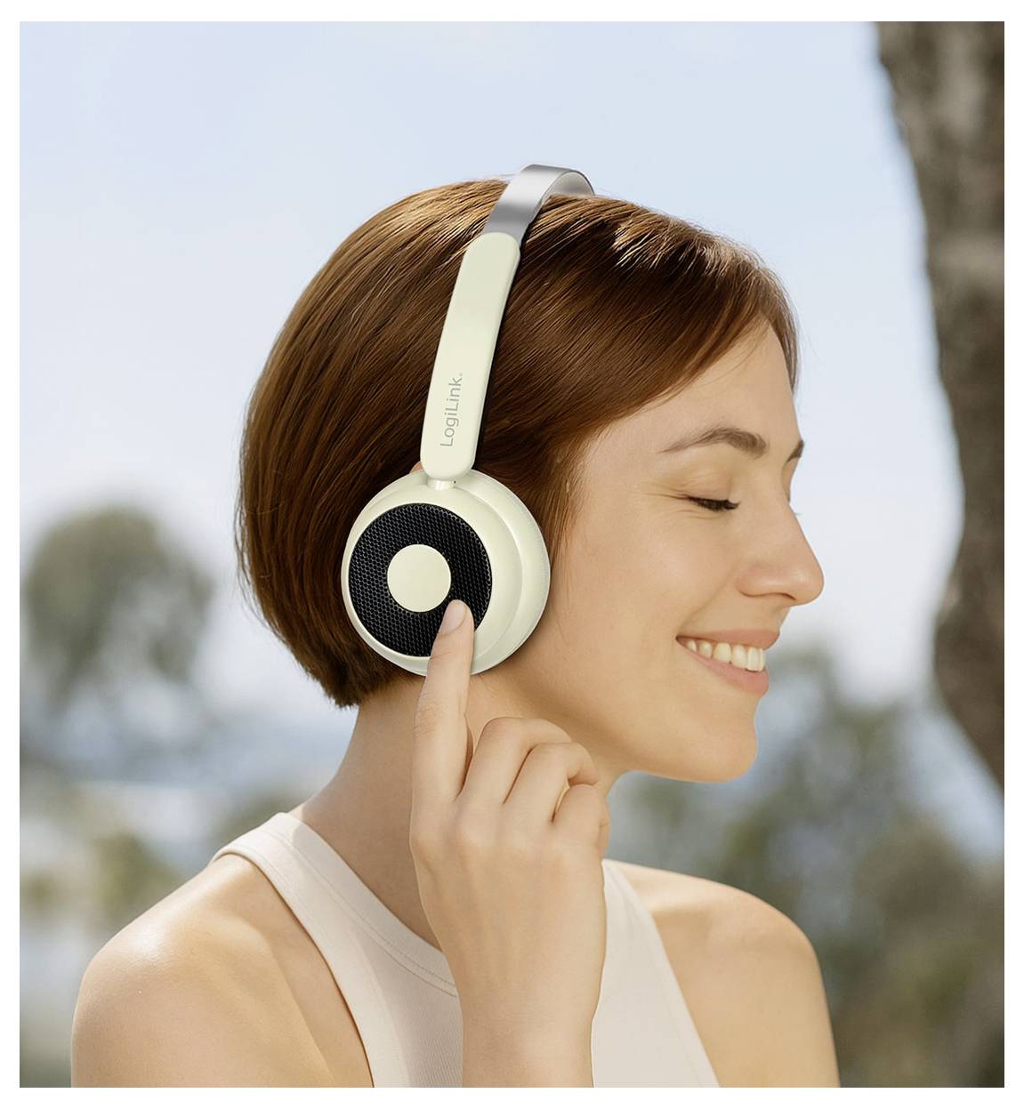 Une personne souriante aux cheveux bruns courts porte des écouteurs circumauraux à l'extérieur, profitant de la musique ou du son, avec des arbres et un ciel bleu en arrière-plan.