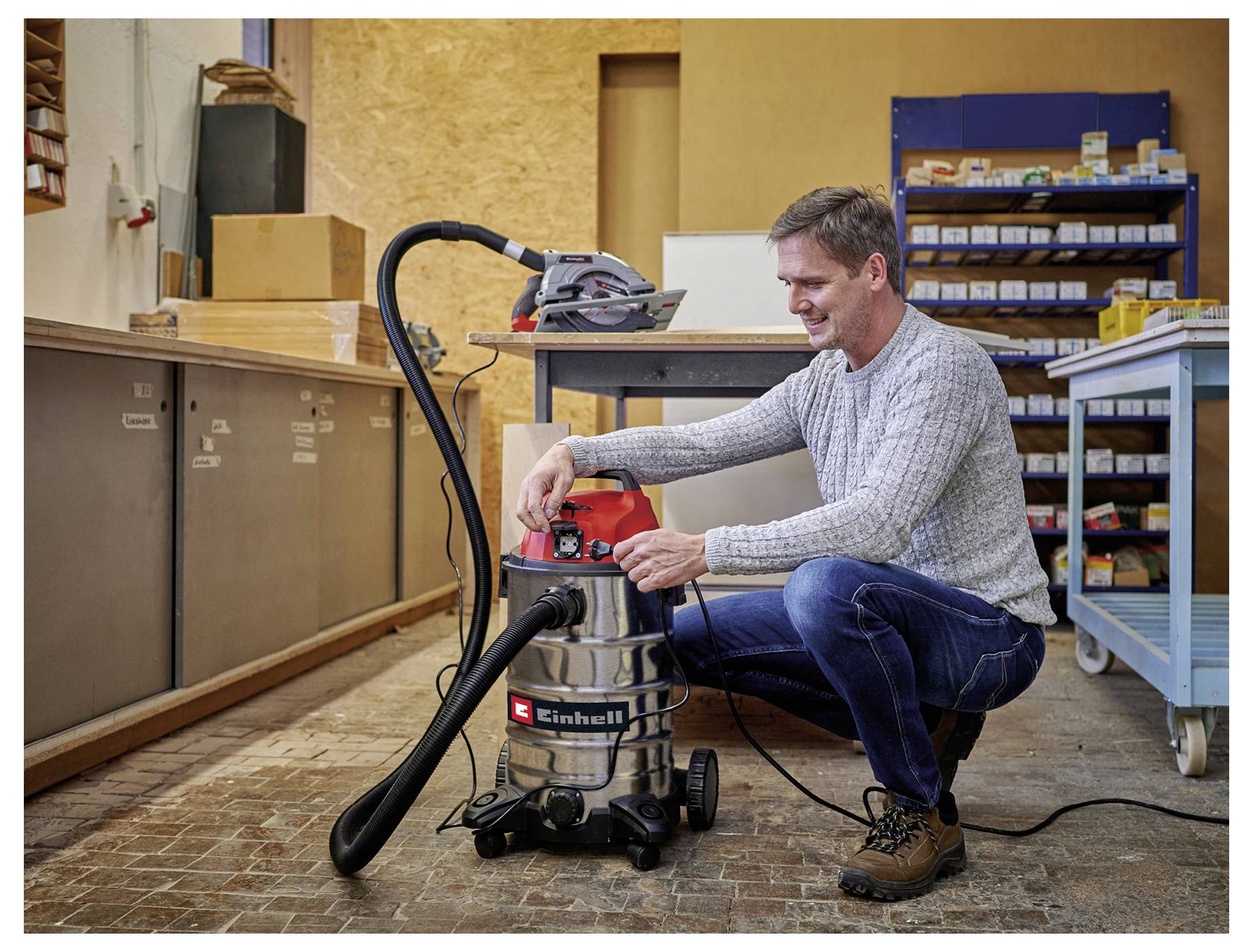 Homme utilisant un aspirateur dans un atelier, entouré d'outils et d'étagères de rangement. Il est en train de régler les paramètres de la machine.
